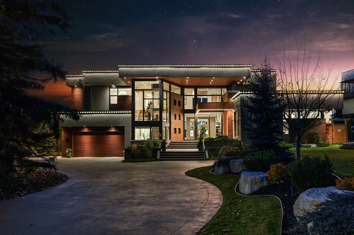 The full front elevation at dusk, its cantilevered flat rooflines edged in light above a double-height glass entry, cedar accents, and a curved drive framed by limestone boulders.