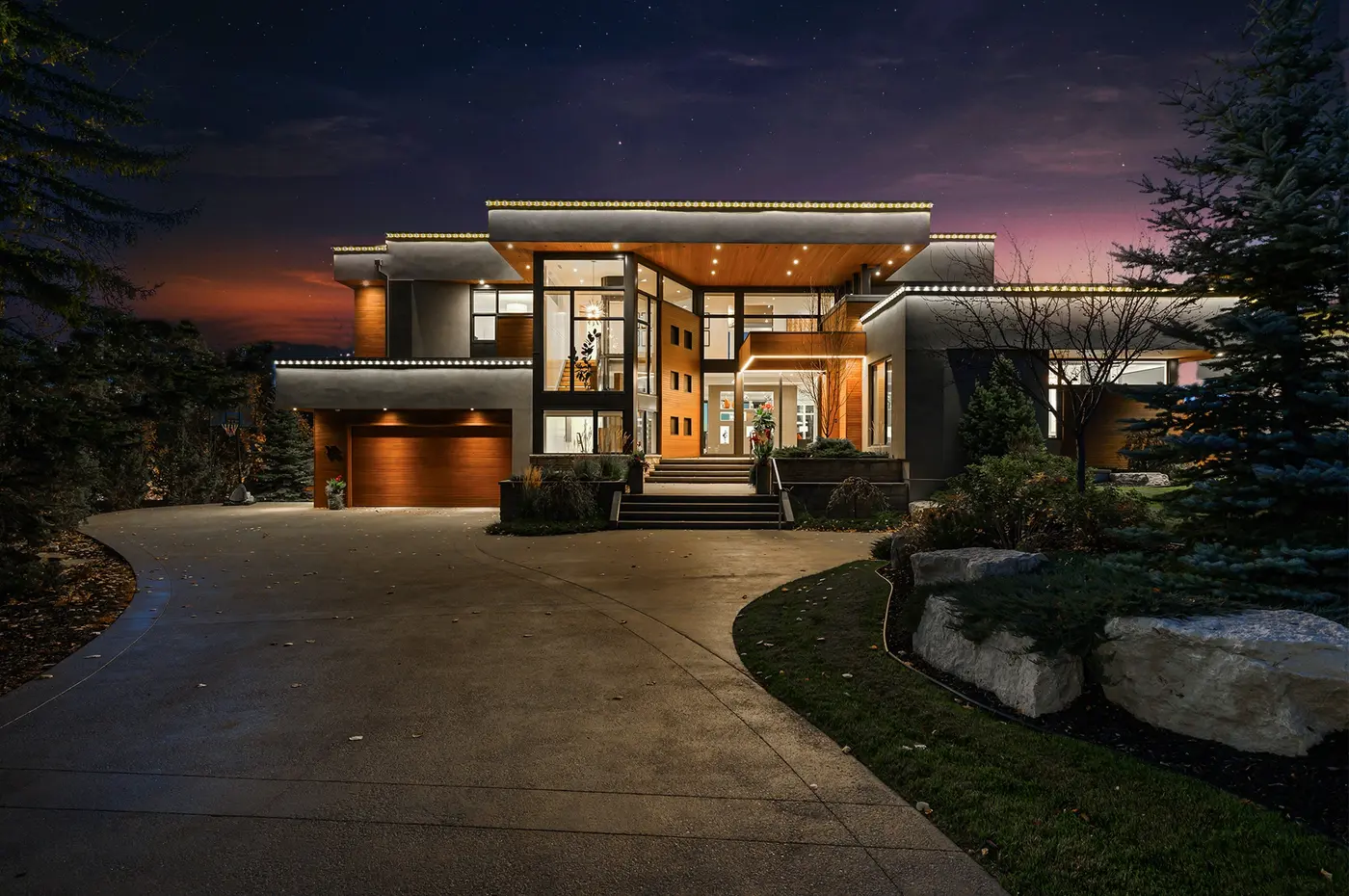A slightly angled twilight view of the facade, where illuminated rooflines float over walls of glass and the warm cedar garage door anchors the composition beneath a violet sky.