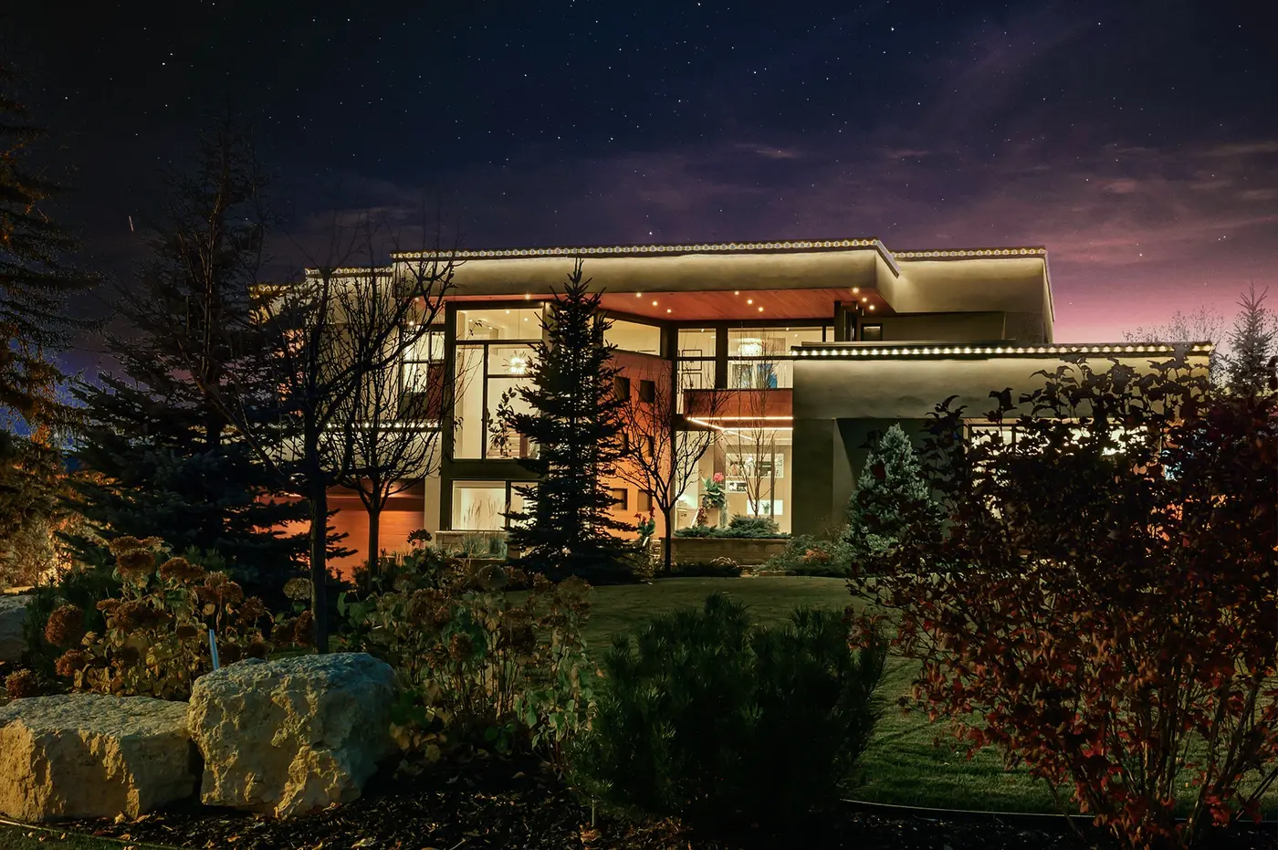 The estate glimpsed through mature evergreens and autumn plantings at night, its lit interiors glowing warmly behind layers of landscaped garden and limestone rock.