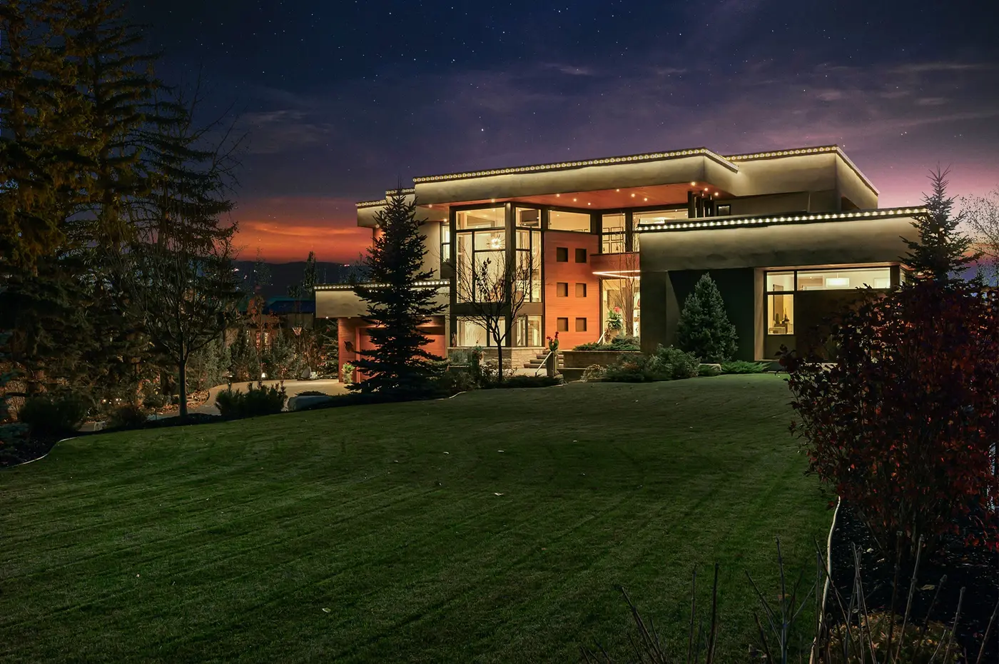 A wide dusk vista across the sloping front lawn as the last orange band of sunset silhouettes distant hills behind the glowing modern silhouette of the home.