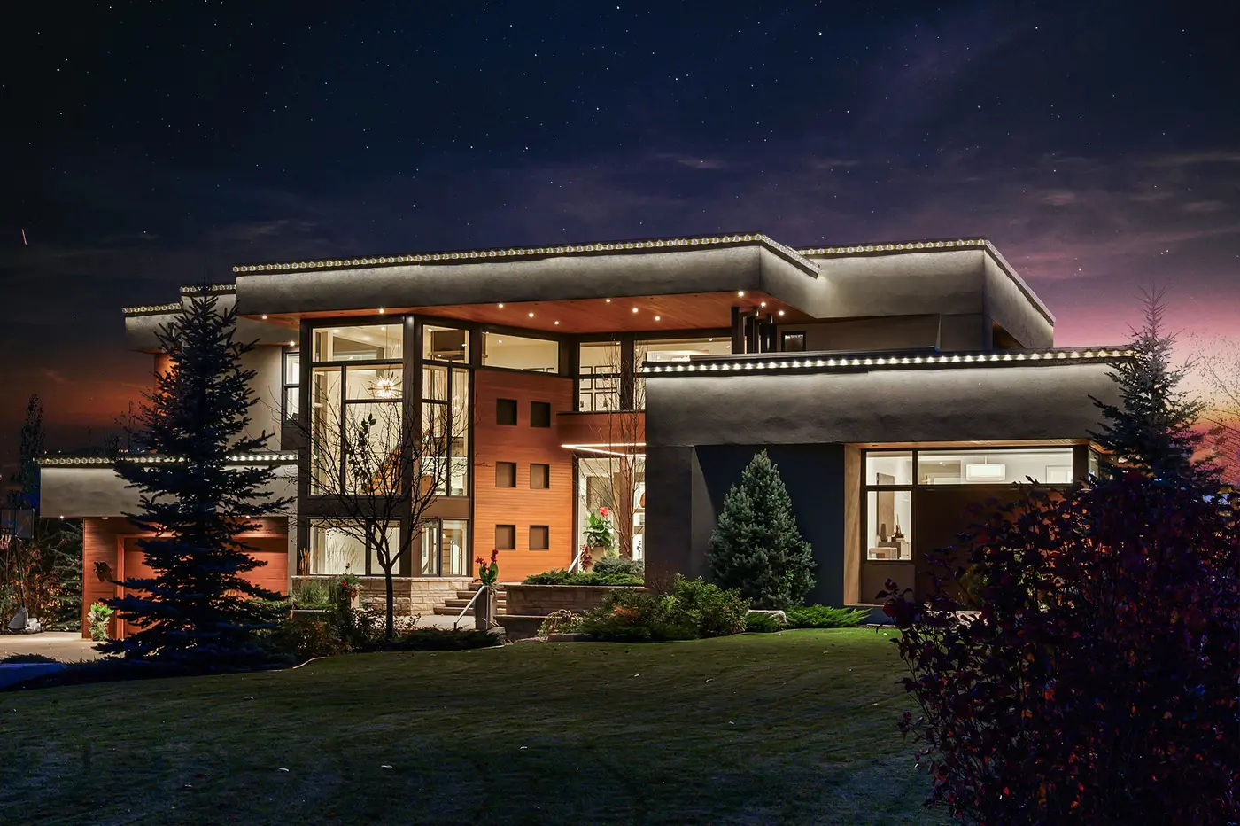 The front elevation seen across the manicured lawn at nightfall, cedar panels punctuated by square windows and a blue spruce standing sentinel beside the stone-clad entry stair.
