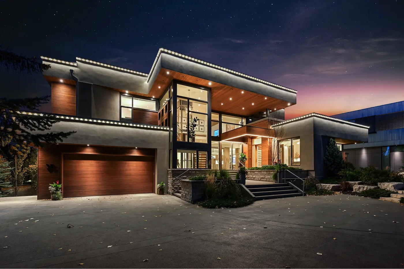 An angled evening study of the front approach, the broad cedar garage door and soffit-lit overhangs leading the eye to the double-height glass foyer and floating entry steps.