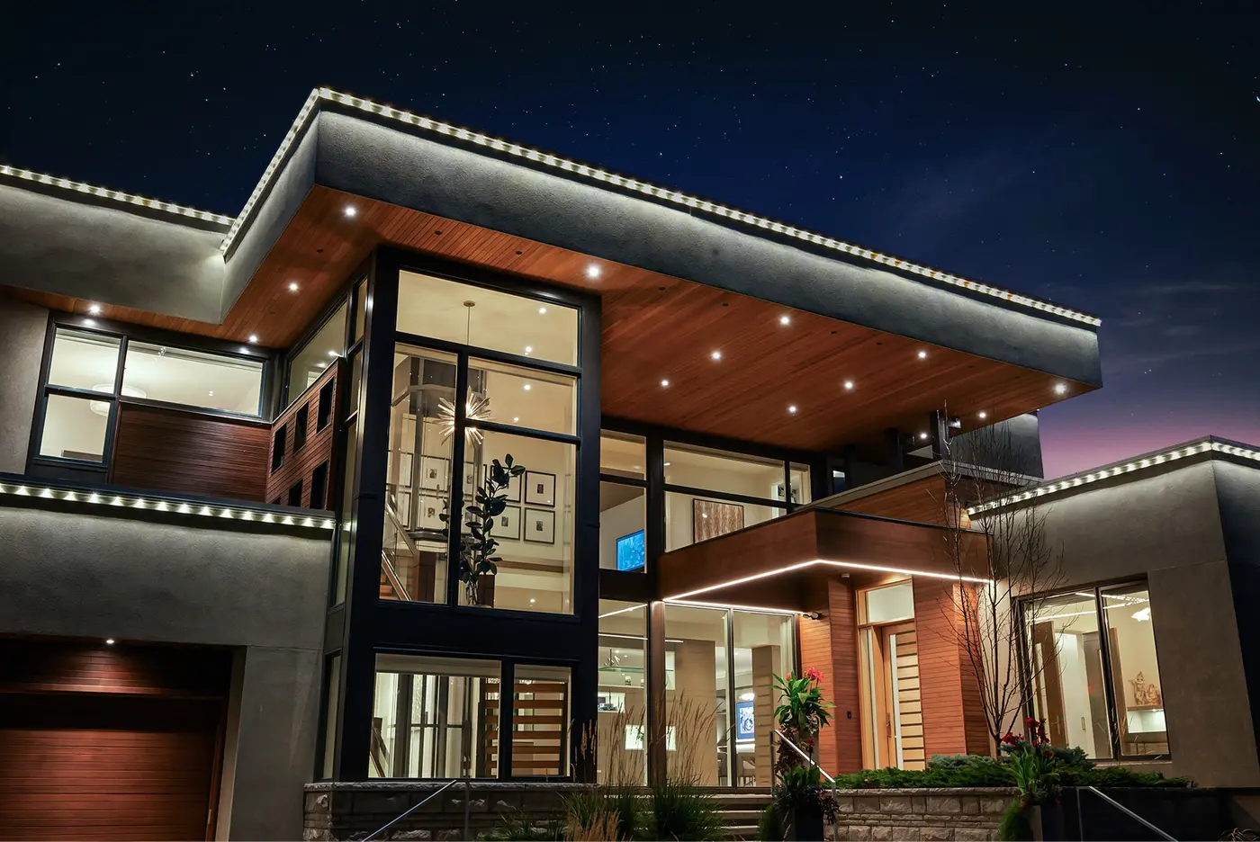 A close twilight portrait of the two-storey glass entry corner, revealing the interior staircase, gallery art wall, and starburst chandelier beneath a cedar-lined canopy studded with pot lights.