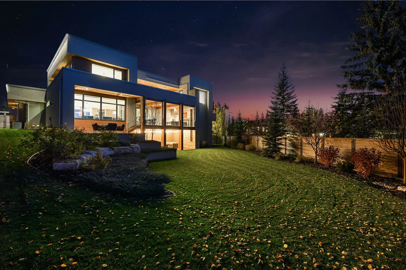 The rear of the residence at night from the lawn, terraced limestone steps and layered plantings climbing to a lit terrace while a horizontal cedar fence encloses the private yard.
