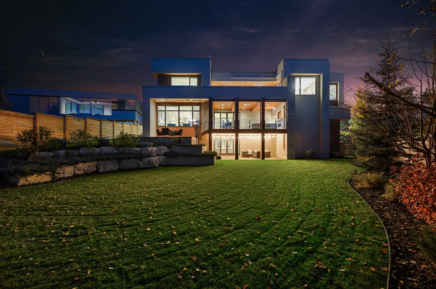 The rear elevation glowing at dusk, a glass-railed main terrace stacked above a string-lit walkout patio, with stacked-stone piers and stucco volumes stepping against the starry sky.