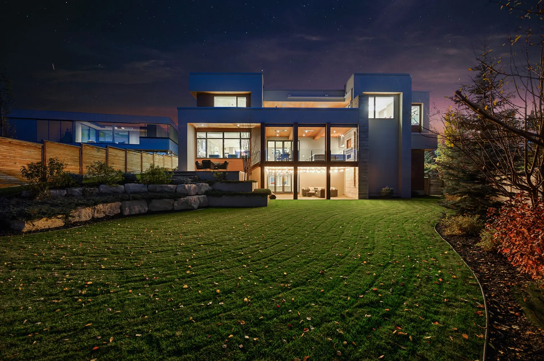 The rear elevation glowing at dusk, a glass-railed main terrace stacked above a string-lit walkout patio, with stacked-stone piers and stucco volumes stepping against the starry sky.