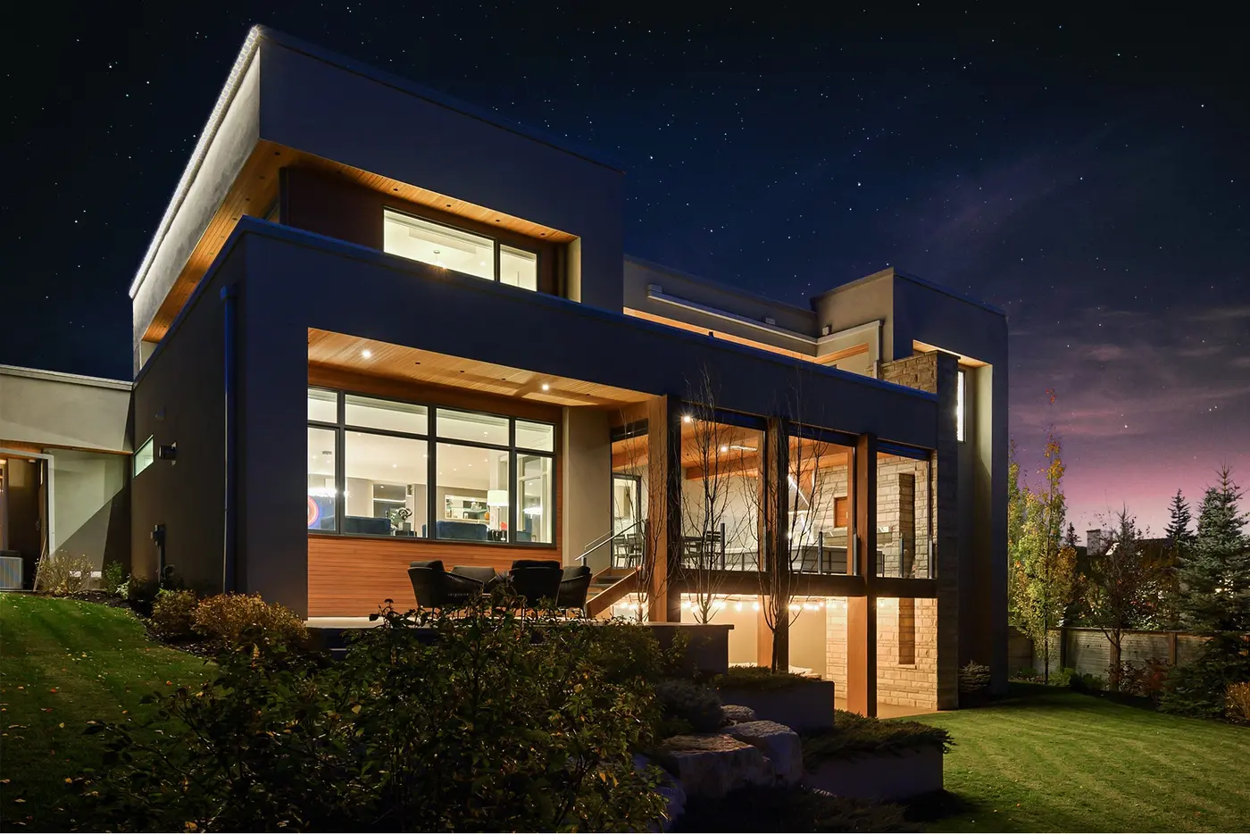A rear corner view at night showing the rooftop deck, halo-lit parapets, and the covered main-level terrace opening to the walkout below through columns of stone and timber.