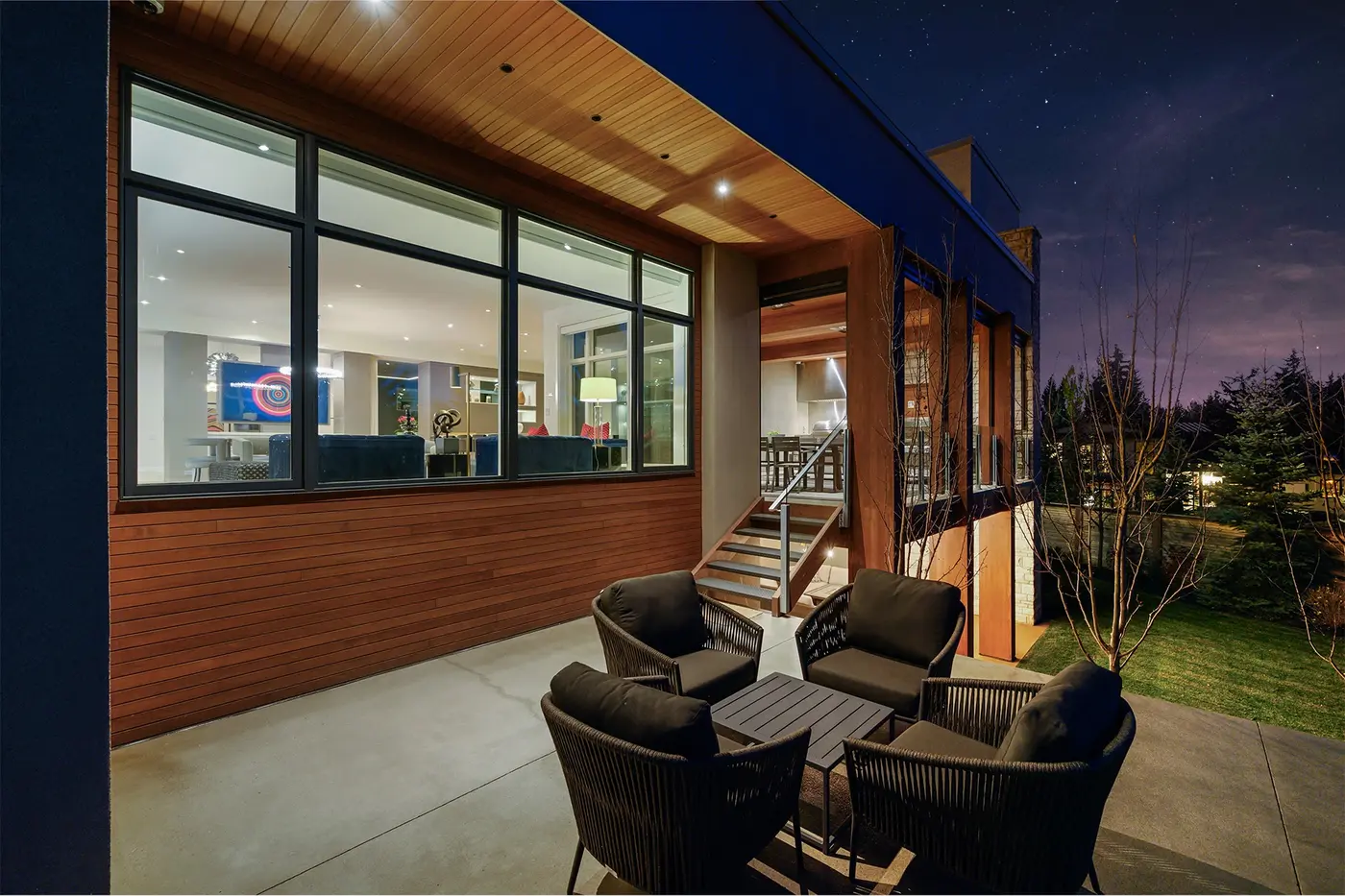The main-level covered terrace at dusk, four woven lounge chairs gathered on polished concrete beside a broad picture window into the family room and stairs descending to the yard.