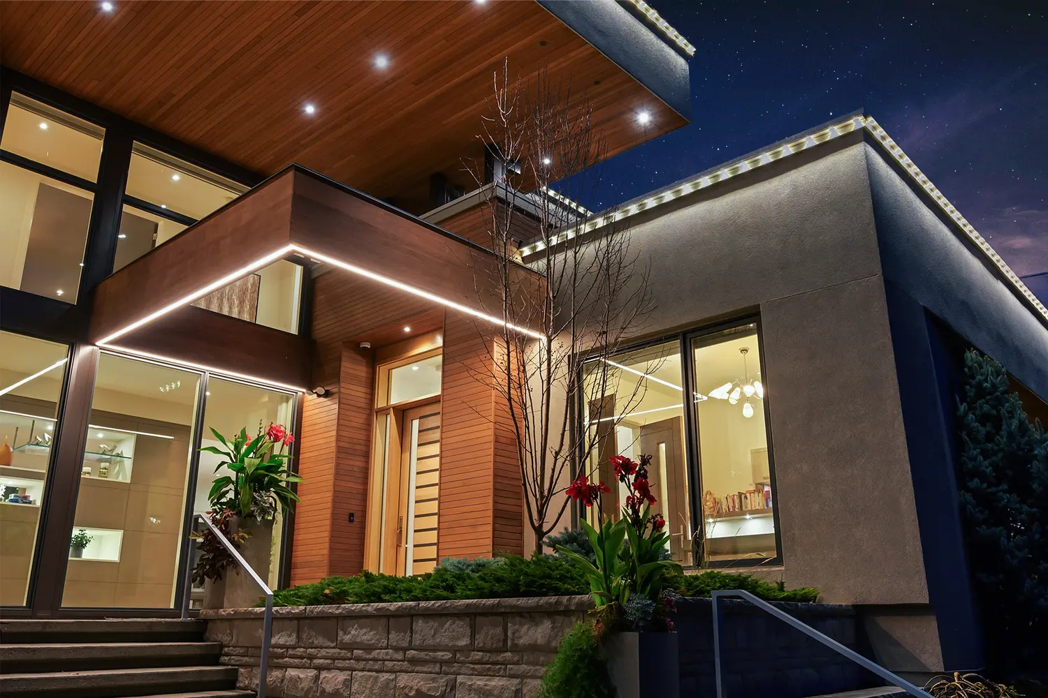 A low evening angle at the front steps where a crisp ribbon of LED light traces the cedar entry canopy above stone planter walls spilling with red cannas.