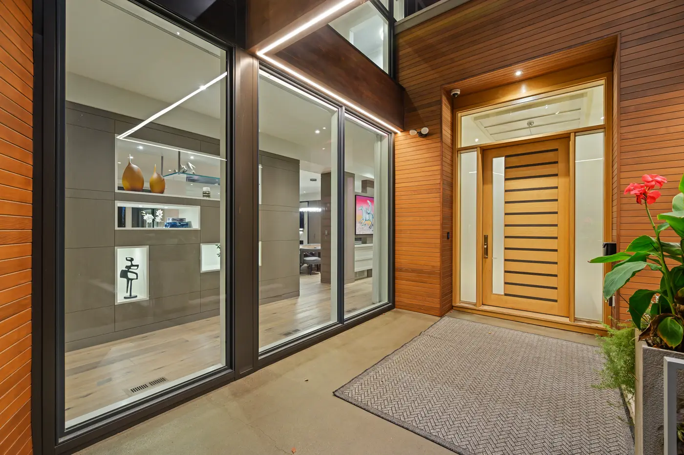 The sheltered entry courtyard at night, the slatted oak door flanked by cedar siding while floor-to-ceiling glass reveals backlit display niches of sculpture and art within.