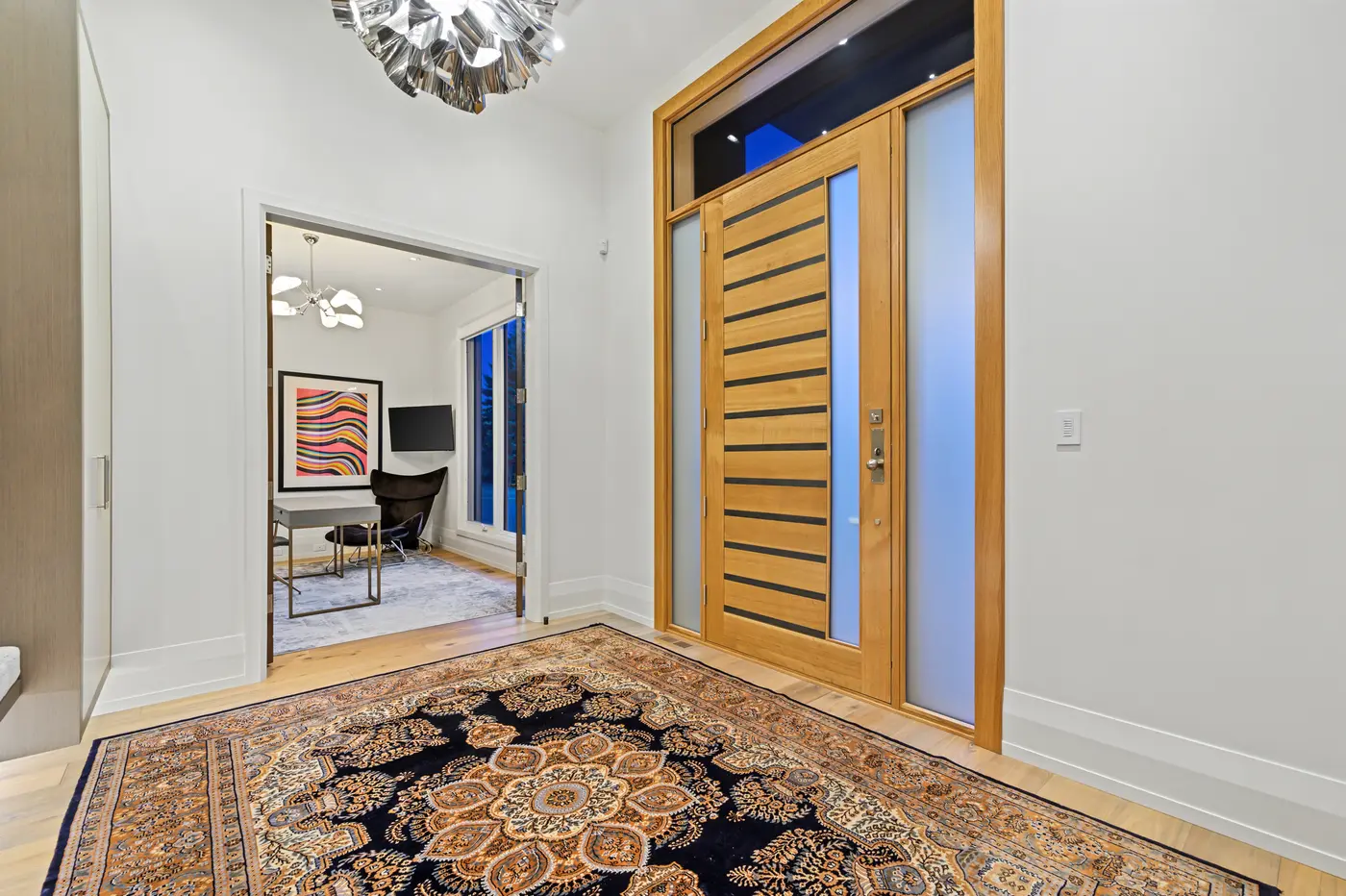The foyer with its slatted white-oak front door and frosted sidelites, a polished-nickel starburst chandelier hovering above a Persian rug and a glimpse of the front office beyond.