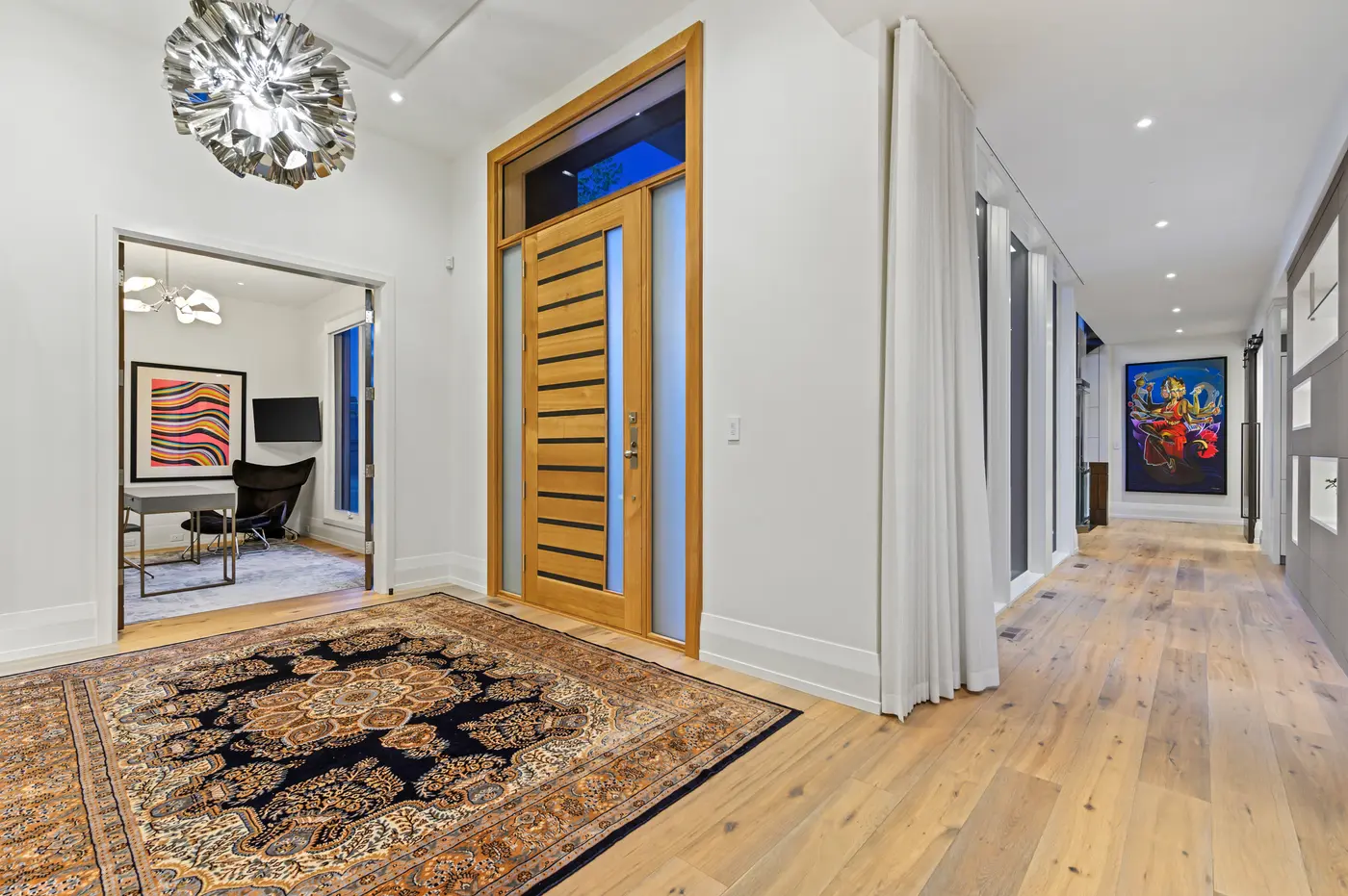 A wide foyer view pairing the oak entry door and Persian rug with a curtained glass gallery hall that draws the eye to a vivid multi-armed deity painting at its end.