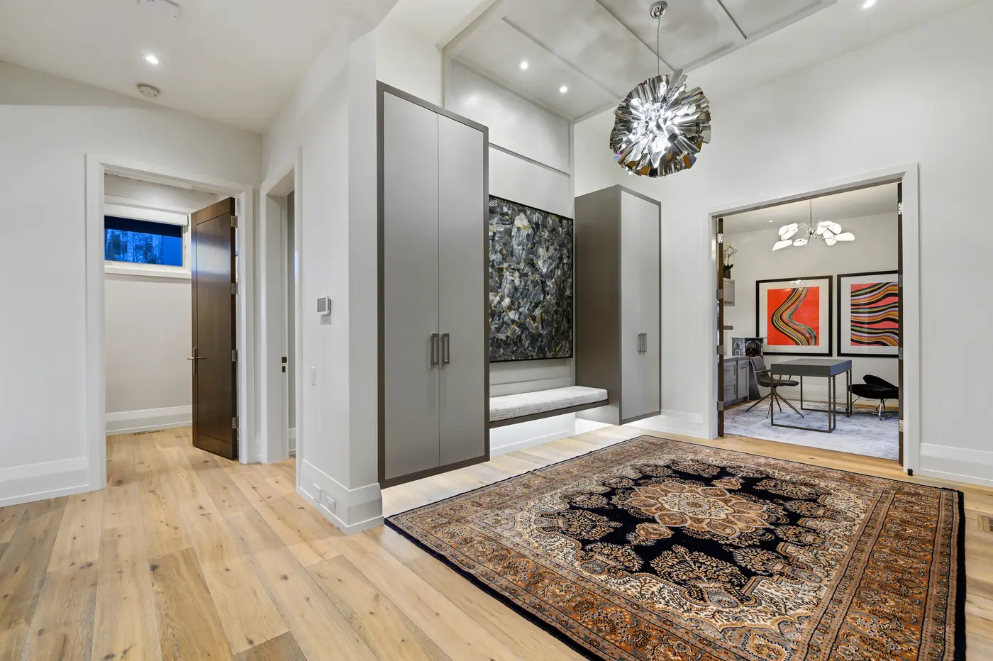 The foyer's dressing area with floating grey wardrobes, an upholstered bench beneath a charcoal-and-gold abstract canvas, and the office's orange wave prints framed through open walnut doors.