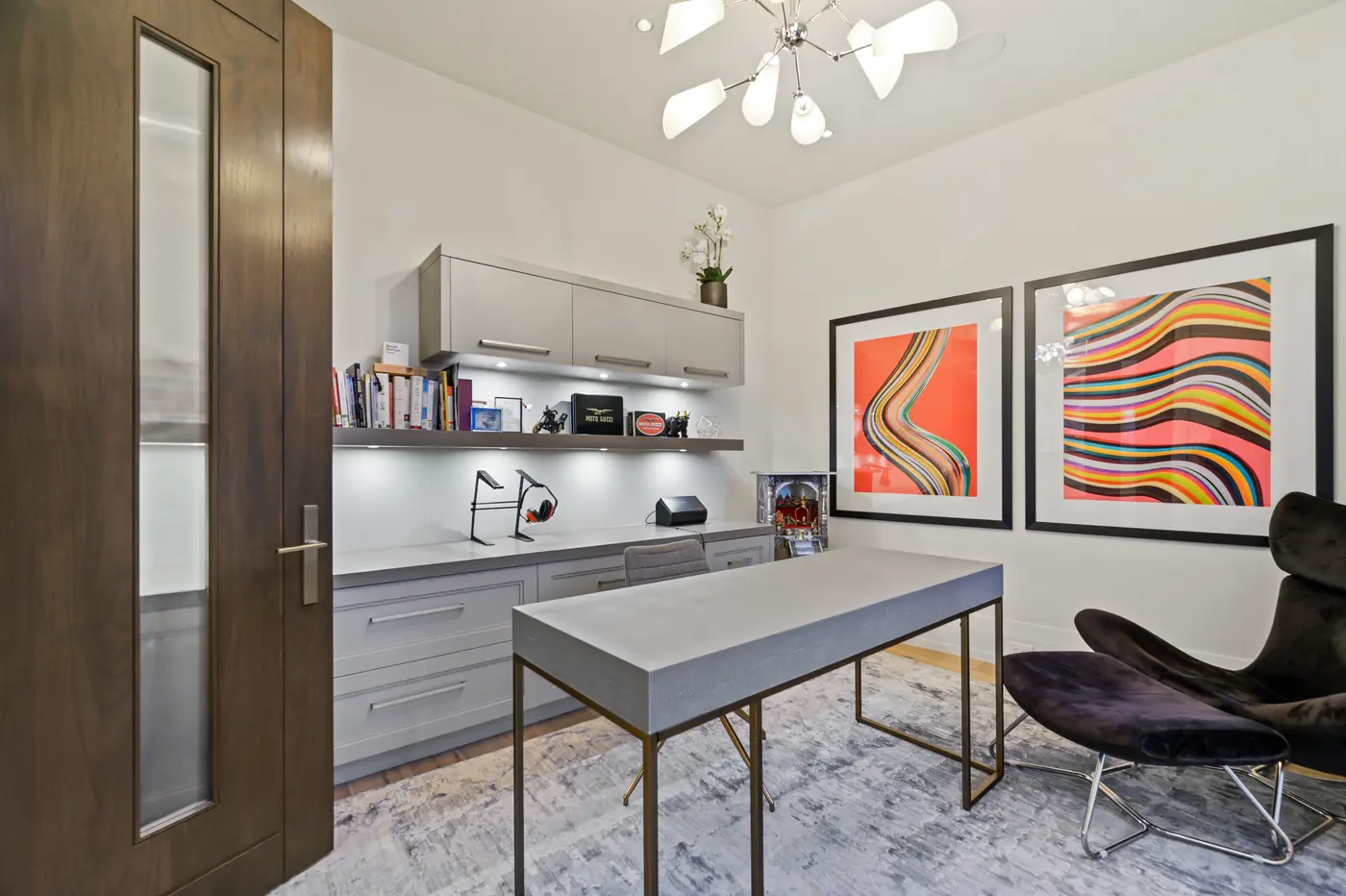 The front office styled with a shagreen-textured desk on a brass base, a velvet wingback chair, under-lit shelving of books and Moto Guzzi memorabilia, and twin psychedelic wave prints.