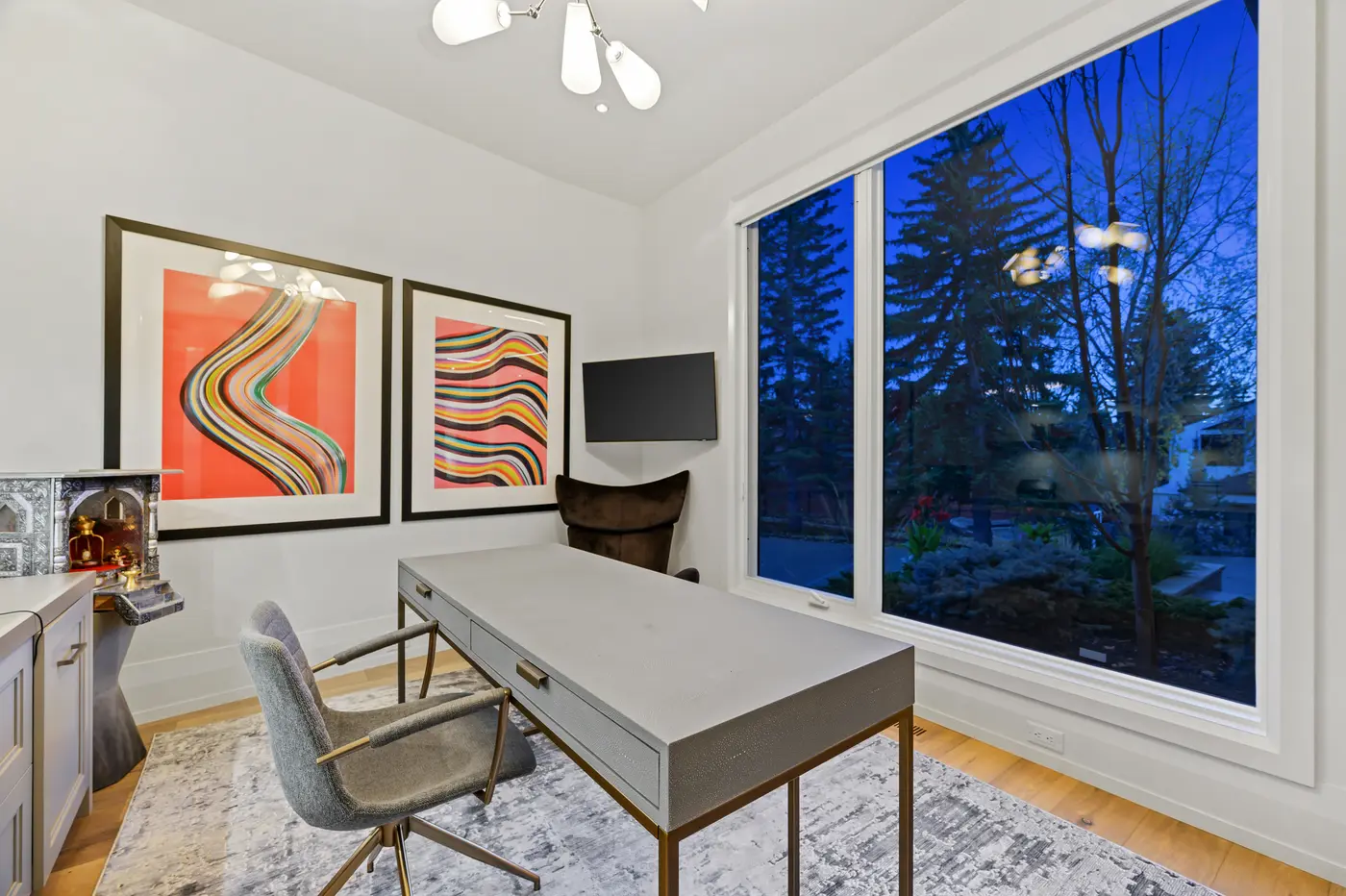 Another angle of the office at dusk, its oversized picture window framing evergreens in deep blue light beside the desk, corner-mounted television, and colourful framed artwork.