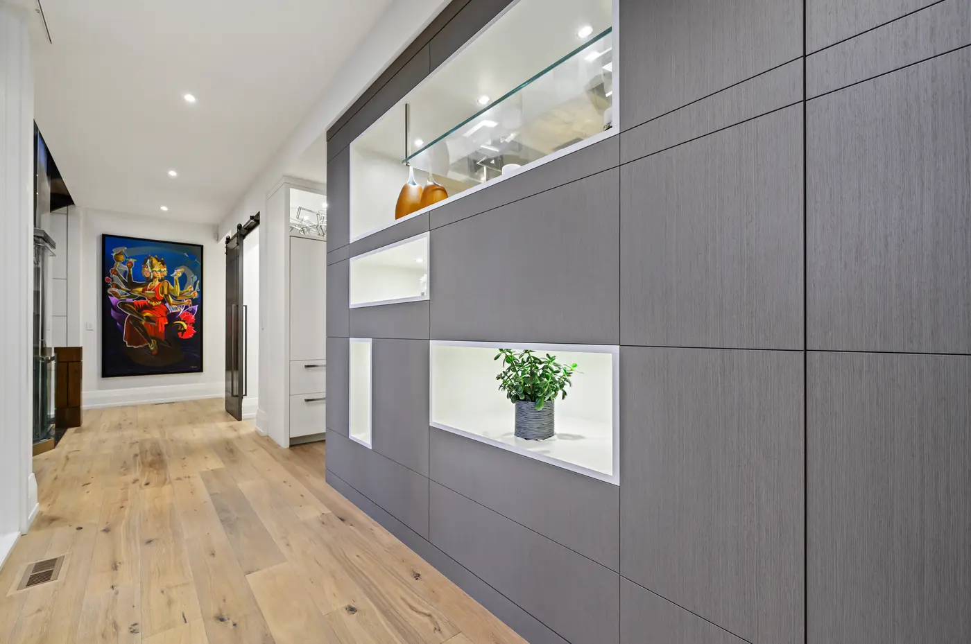 A hallway feature wall of rift-cut grey oak inset with backlit niches holding amber glass and a jade plant, leading toward a barn door and the home's vivid deity painting.