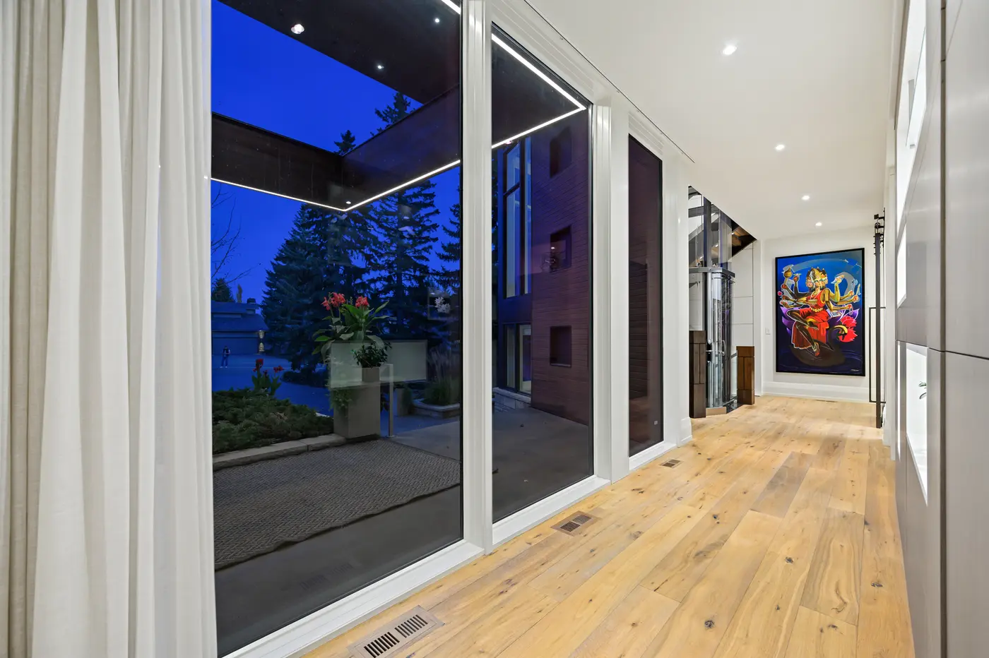 The glass-walled gallery hall at dusk, sheer drapery and entry-courtyard views on one side and the cylindrical glass elevator and bold crimson artwork glowing at its end.