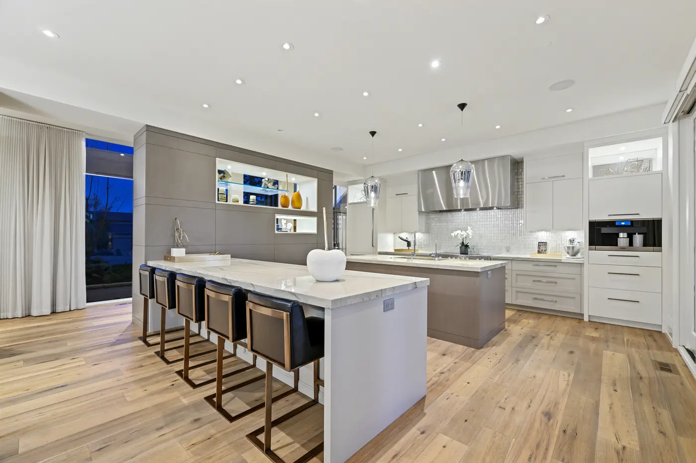 The double-island kitchen with thick-mitred marble counters, black leather and brass stools, a sculptural stainless hood over shimmering mosaic tile, and a built-in Miele coffee centre.