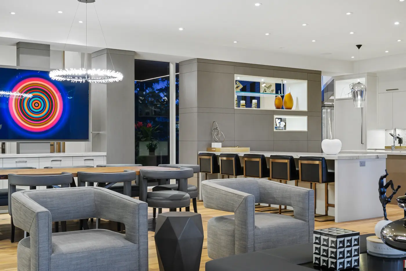 The open entertaining space where dining chairs and bar seating layer toward the kitchen, a halo chandelier hovering and a concentric-ring artwork glowing on the media wall.