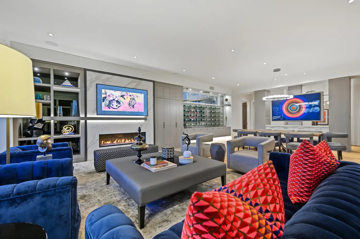 The great room dressed in navy channel-tufted velvet and coral geometric pillows, a linear fire flickering beneath framed TV art in bookmatched marble with the lit wine wall beyond.