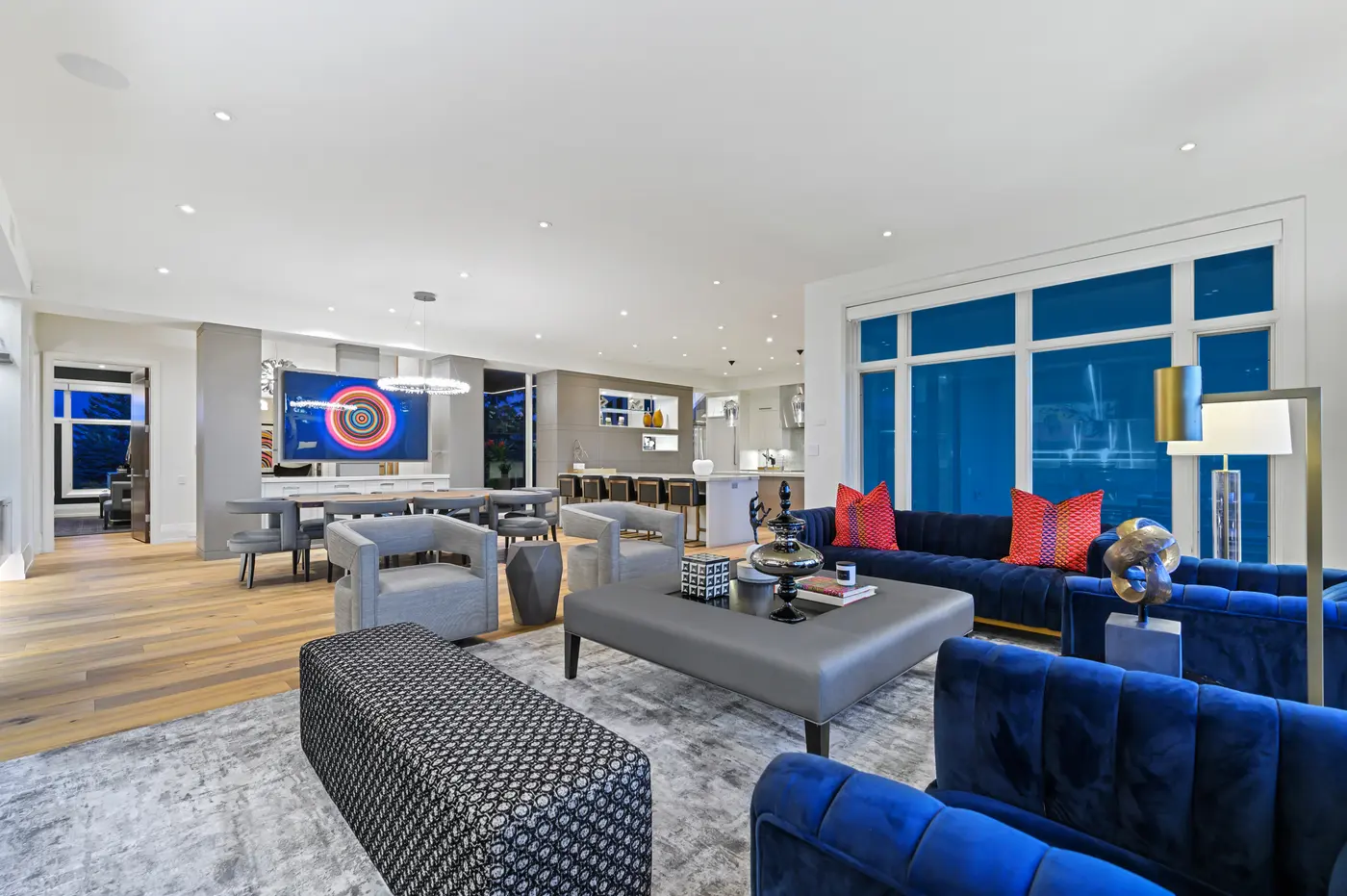 A reverse view across the living room's leather ottoman and patterned bench toward the dining table and kitchen, twilight deepening in the wall of windows.