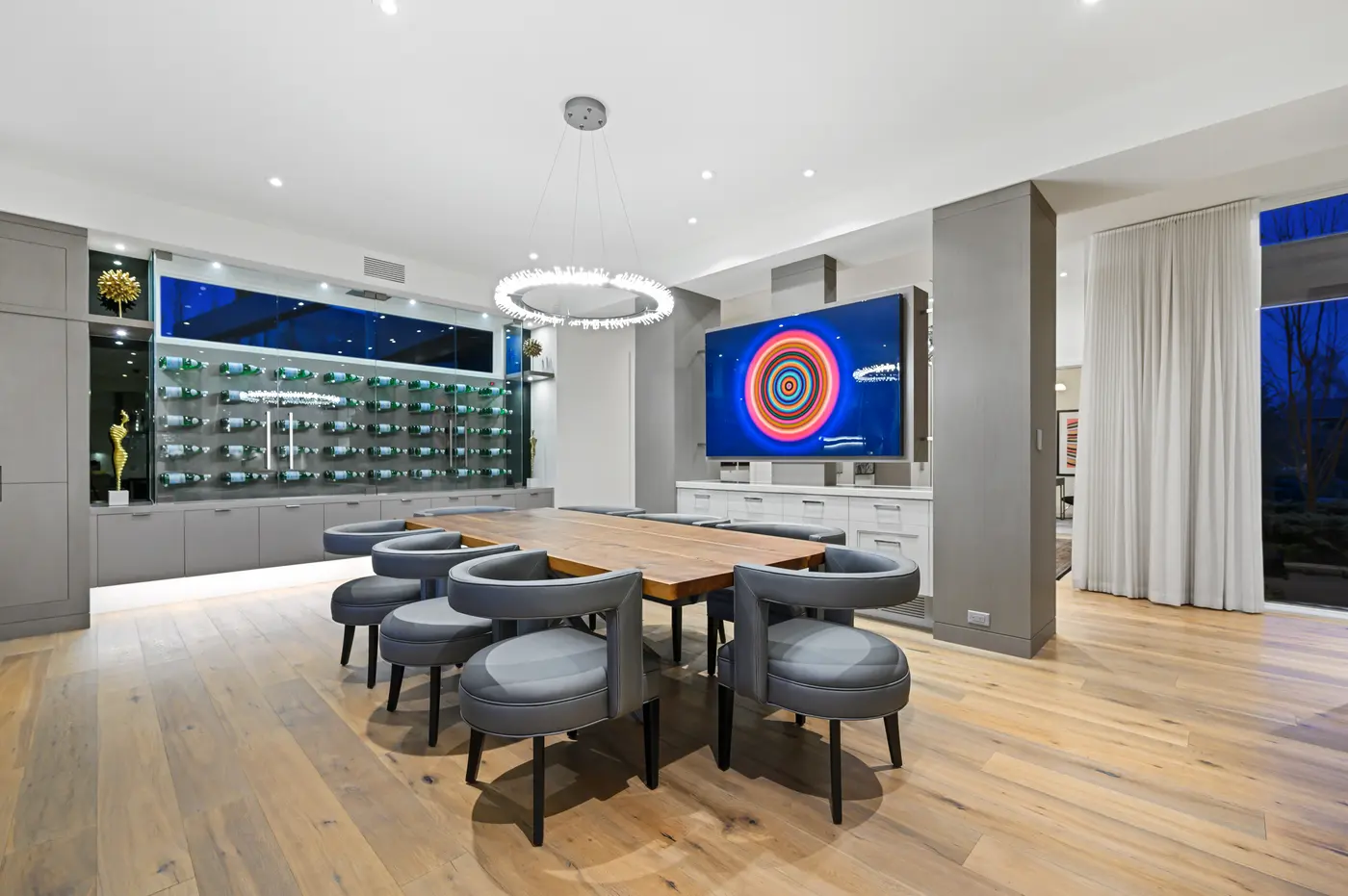 The dining room's live-edge walnut table ringed by grey leather barrel chairs beneath a crystal-fringed halo chandelier, facing a glass-enclosed, backlit wall of bottle display.