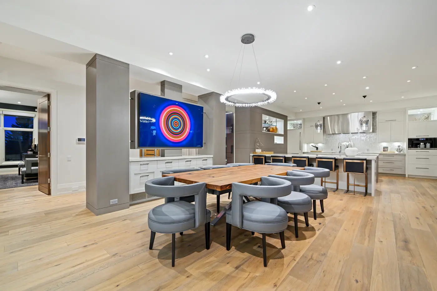 The dining table viewed toward the kitchen, its ring chandelier centred over walnut planks while the rotating media panel's concentric-circle artwork adds a jolt of colour.