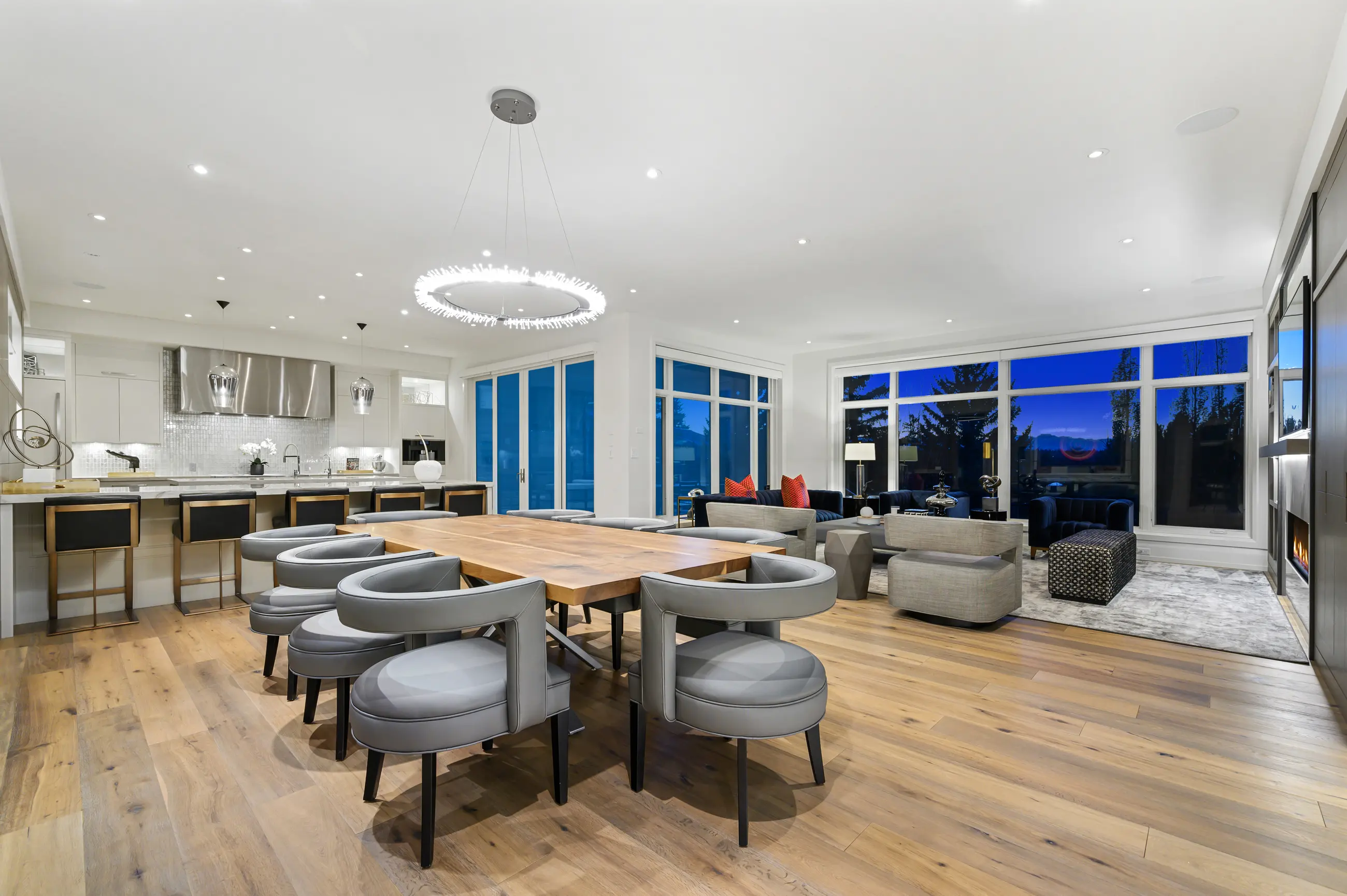 A wide evening panorama of the entertaining level, dining chairs and brass-based stools in the foreground and the navy-and-coral living room glowing against indigo windows beyond.