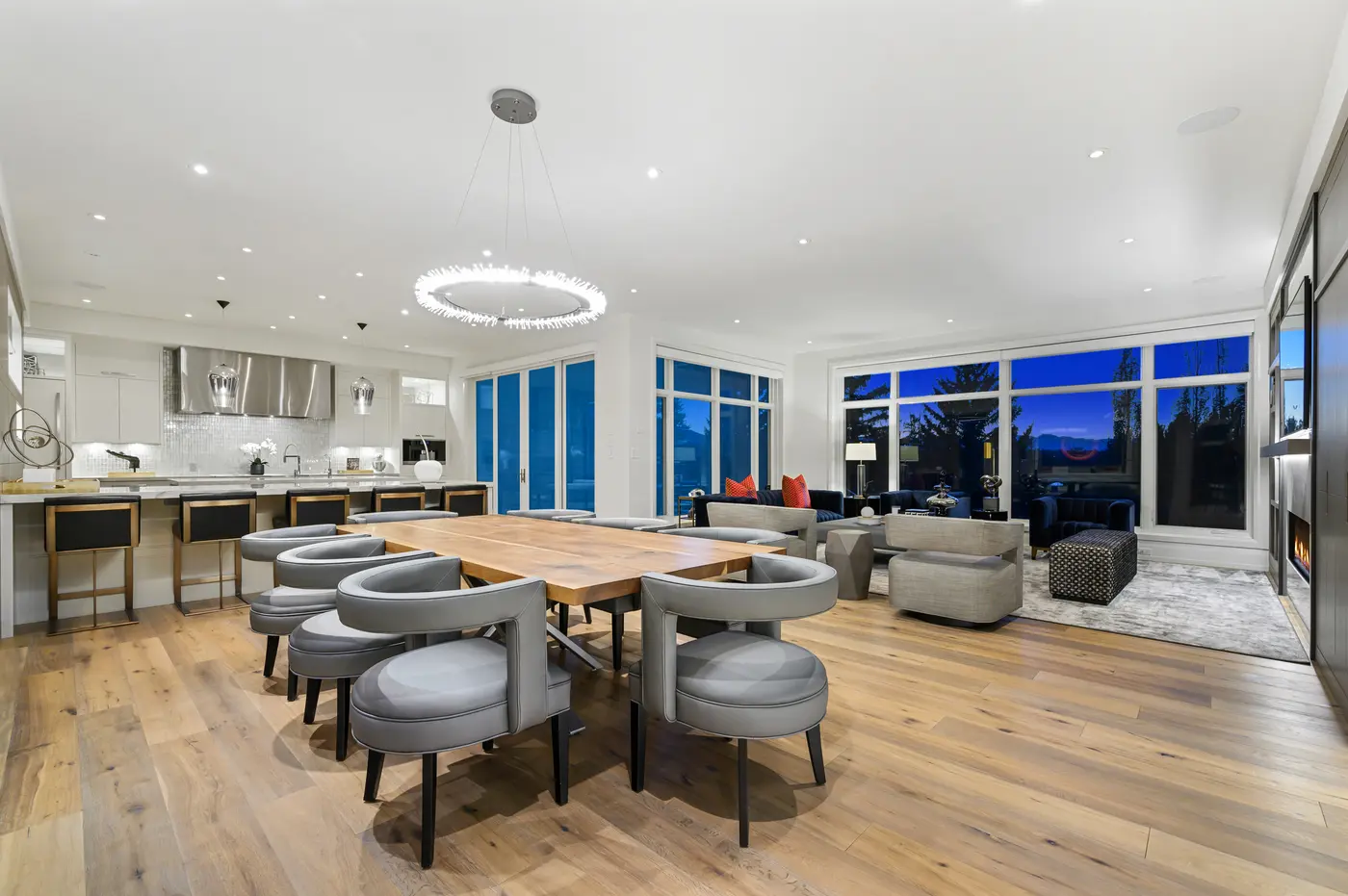 A wide evening panorama of the entertaining level, dining chairs and brass-based stools in the foreground and the navy-and-coral living room glowing against indigo windows beyond.