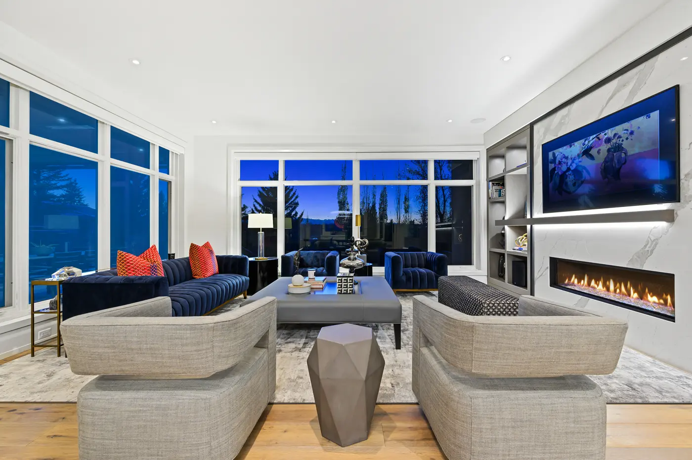 The living room at blue hour, twin navy sofas and swivel chairs arranged around the grey leather ottoman as the window wall dissolves into silhouetted poplars and mountain dusk.