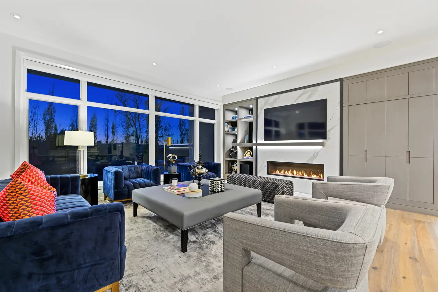An angled view of the great room pairing channel-stitched navy velvet and coral pillows with the marble fireplace, lit display shelving, and a full wall of taupe oak cabinetry.