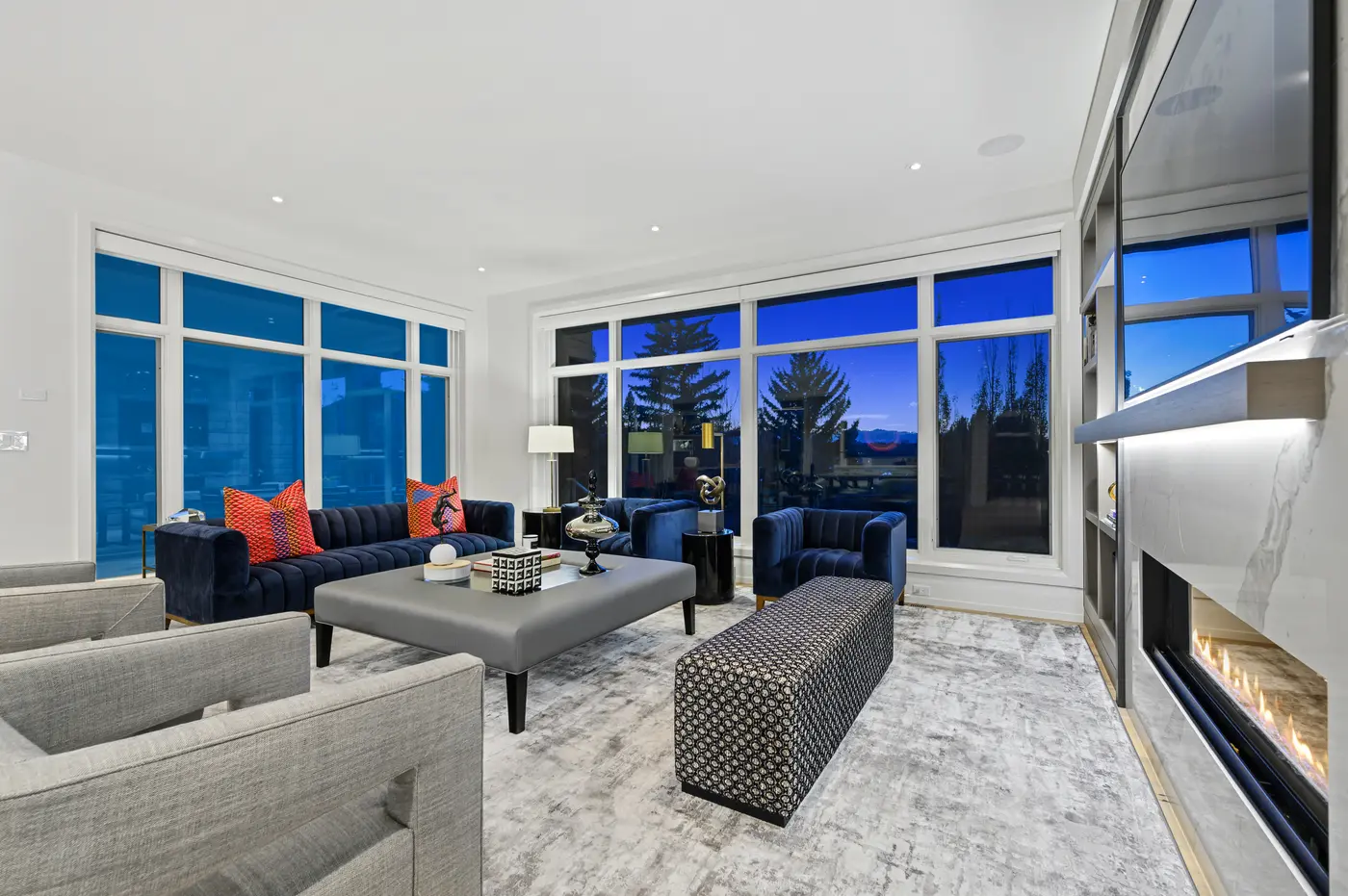 From the corner armchairs, the living room stretches past the patterned bench and linear fireplace toward deep blue windows, layering greys, navy, and warm oak floors.