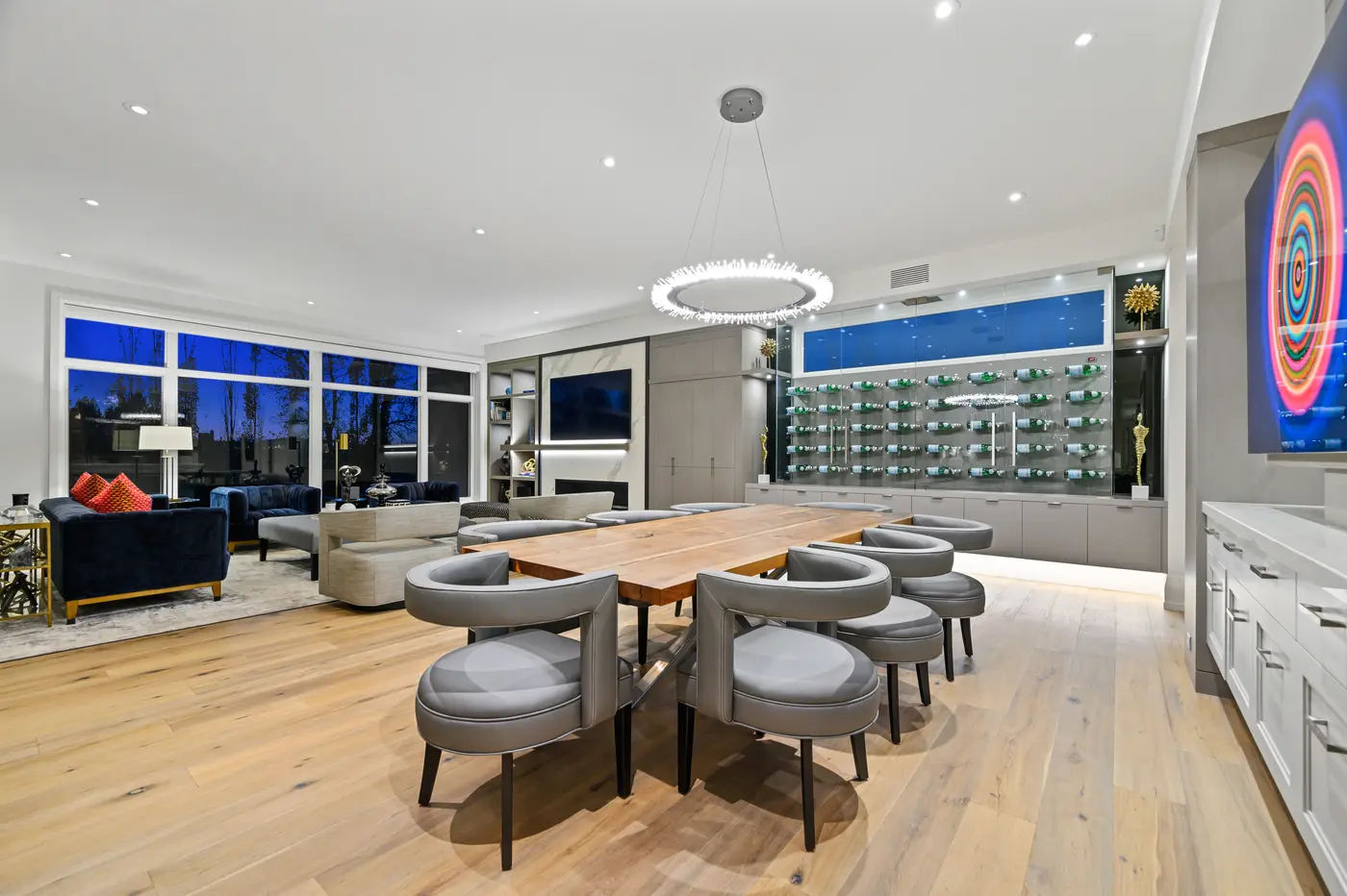The dining space seen from the wine display side, grey barrel chairs circling the walnut table with the lounge's navy sofas and glowing sunset windows in the distance.