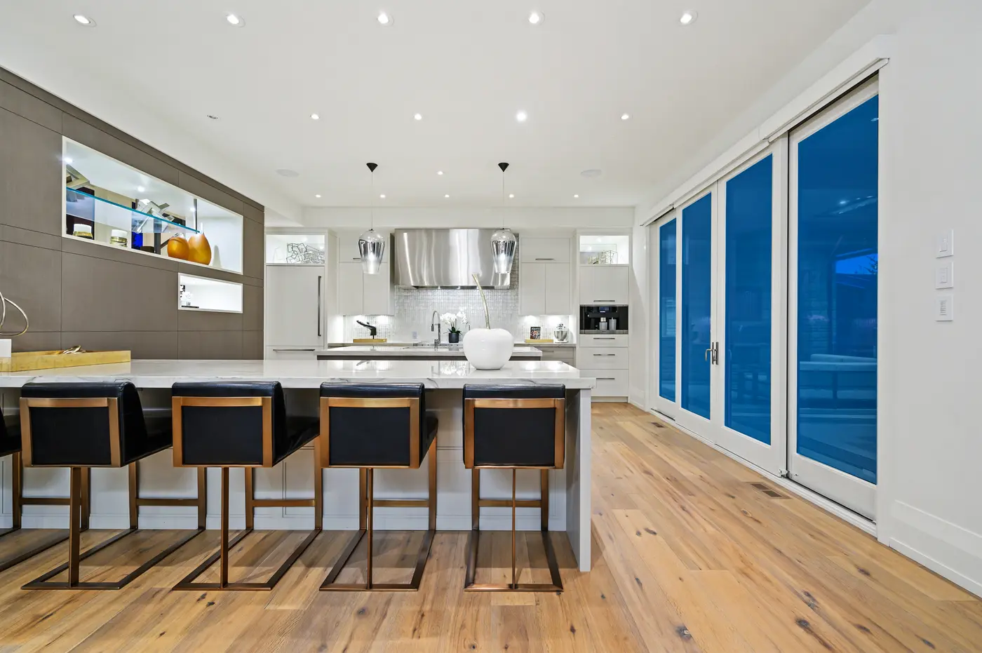 The breakfast bar's black leather and bronze stools lined along marble, with a wall of white-framed glass doors opening the kitchen to the covered terrace at dusk.
