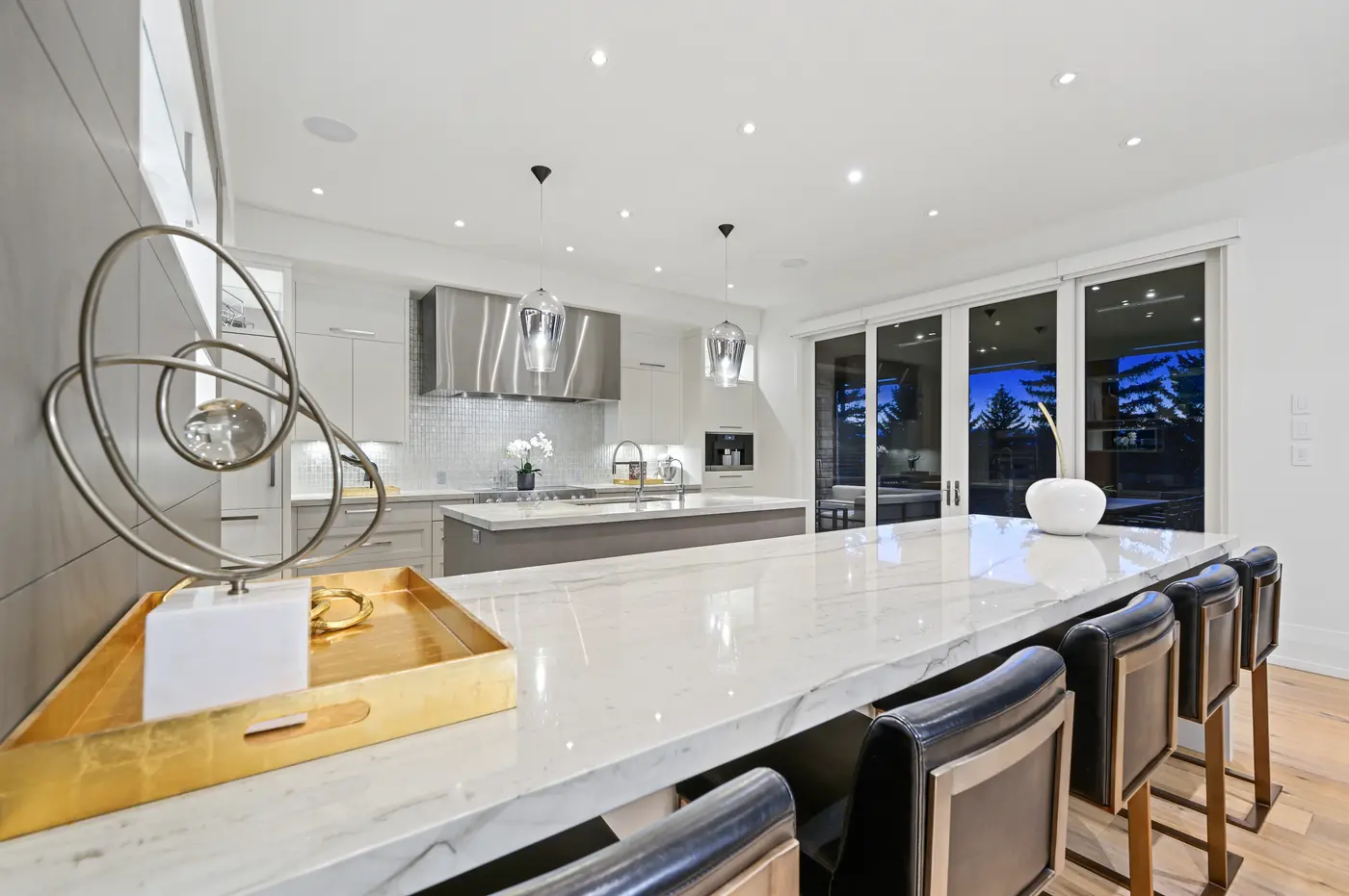 A counter-level study of the honed marble breakfast bar, gold-leaf tray and wire sculpture in the foreground with the orchid-topped prep island and stainless hood beyond.