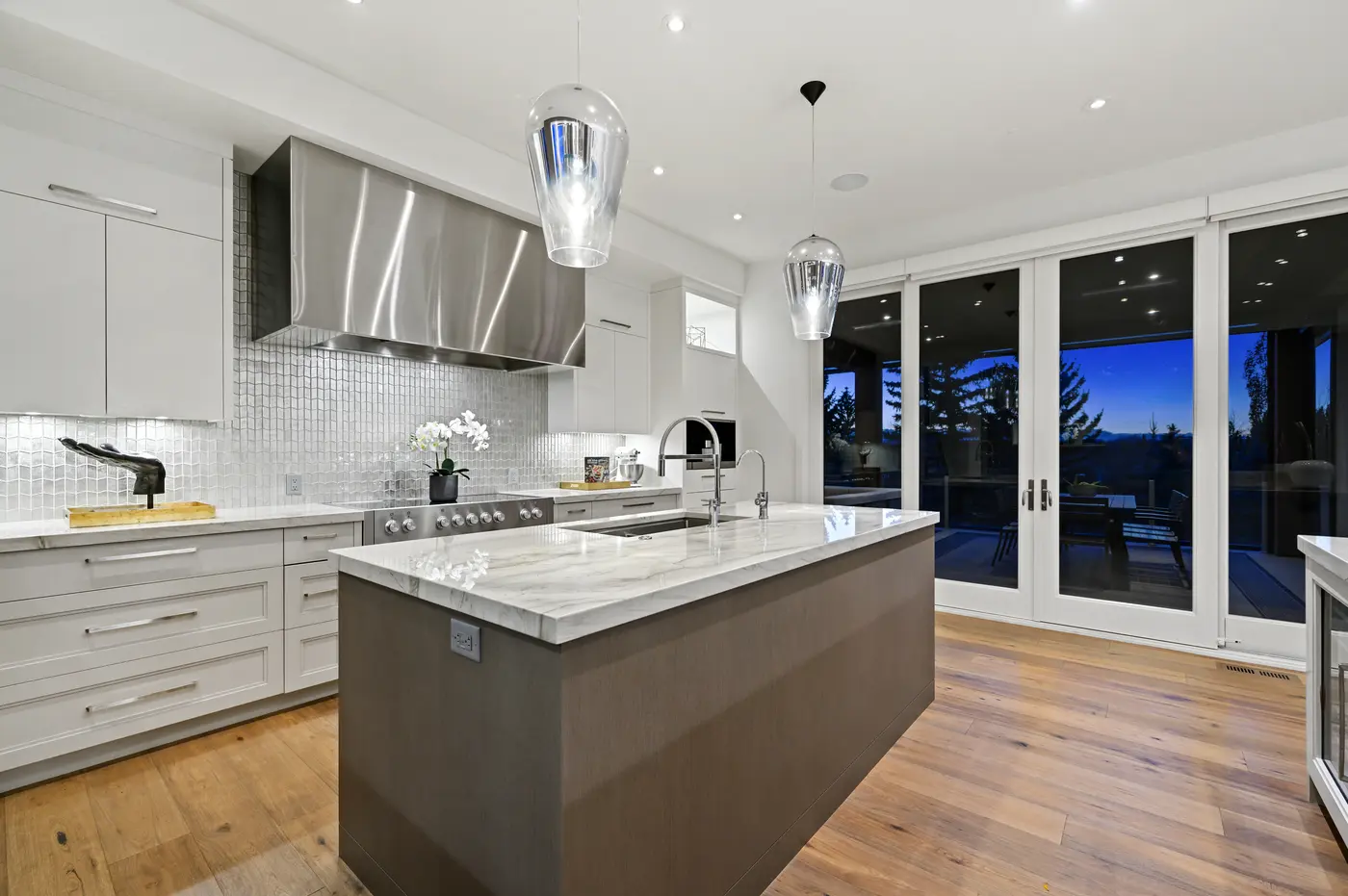 The working side of the kitchen, mercury-glass pendants hanging above the grey-oak prep island and its marble slab, with glass doors framing the twilight terrace beyond.