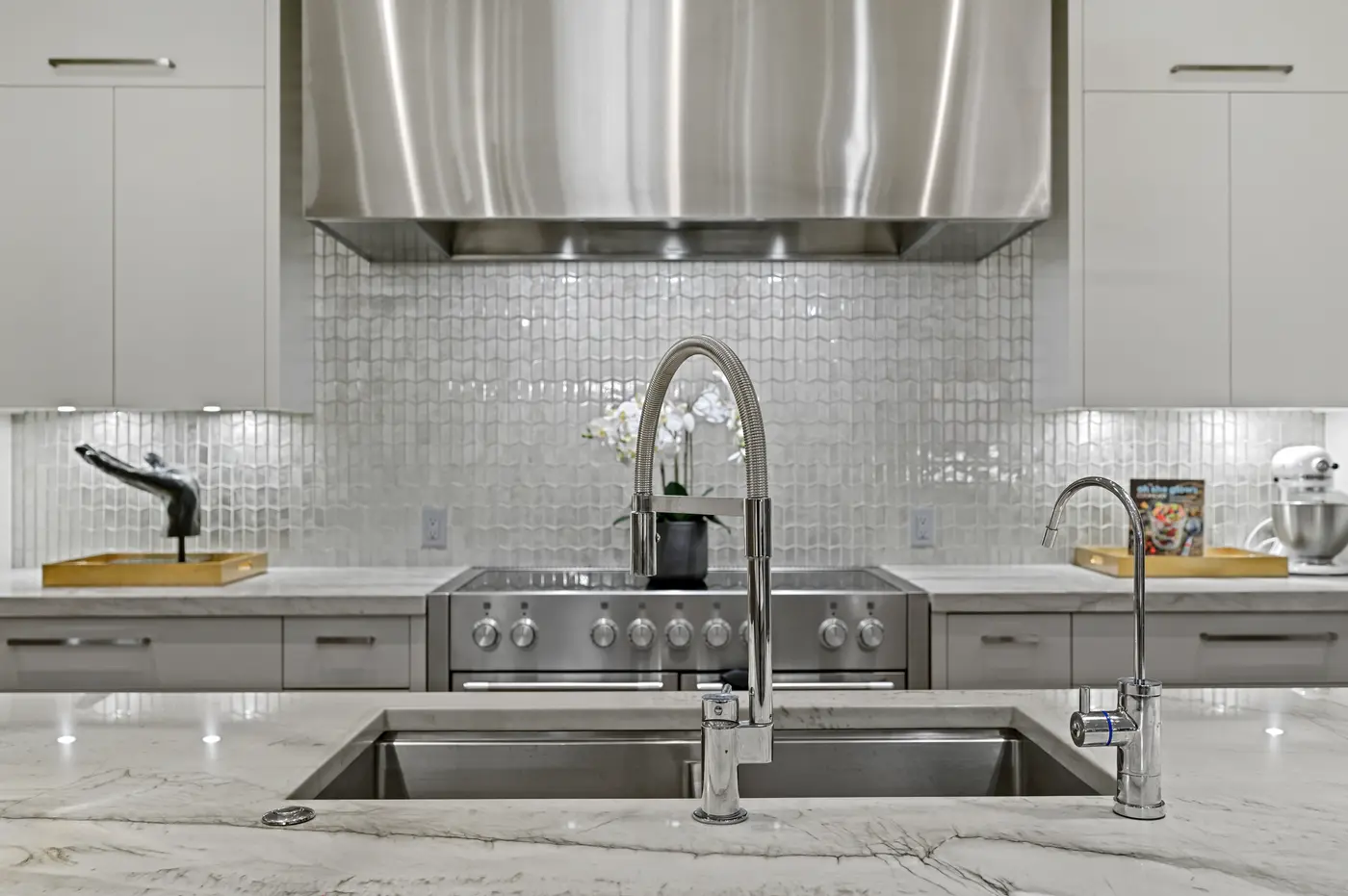 A detail across the island sink, its coiled professional faucet and filtered-water tap standing against the stainless range and pearlescent picket-tile backsplash.