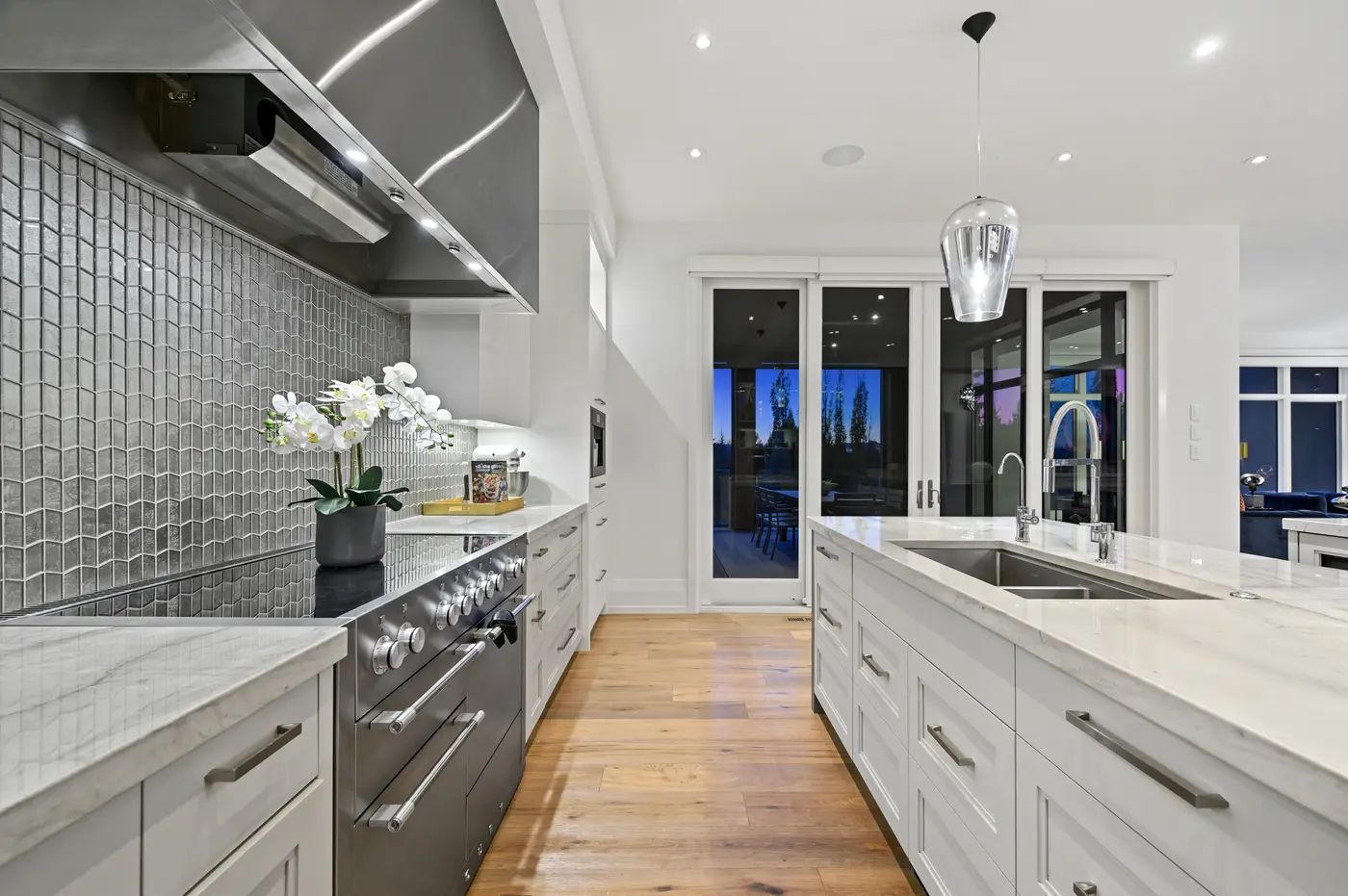 A galley perspective down the kitchen, professional stainless range and hood set into shimmering picket-mosaic tile opposite the long island's twin faucets and drawer banks.