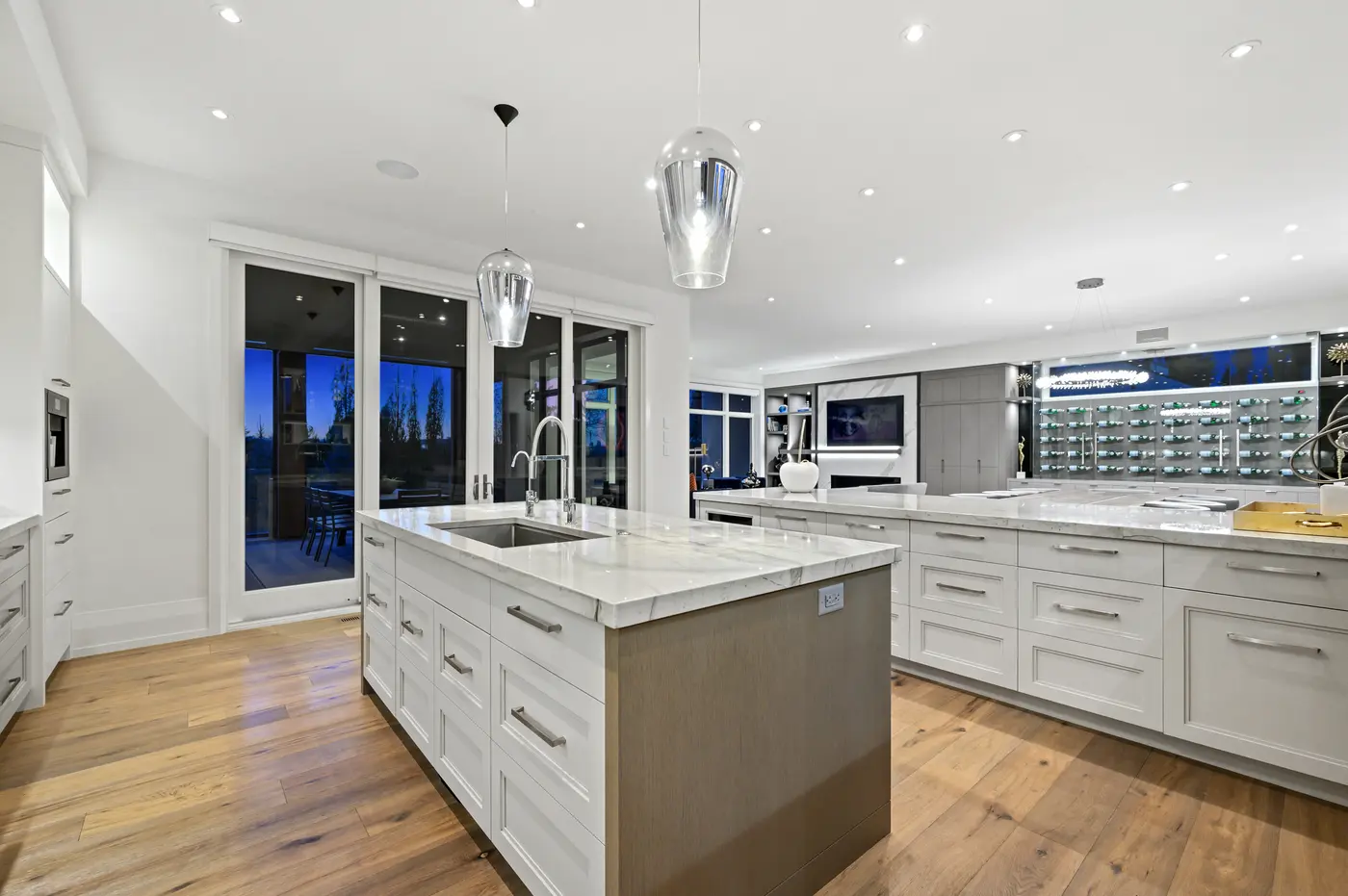 Both islands seen from the pantry corridor, an under-counter wine fridge tucked into the marble bar as the backlit bottle wall glows across the dining room.
