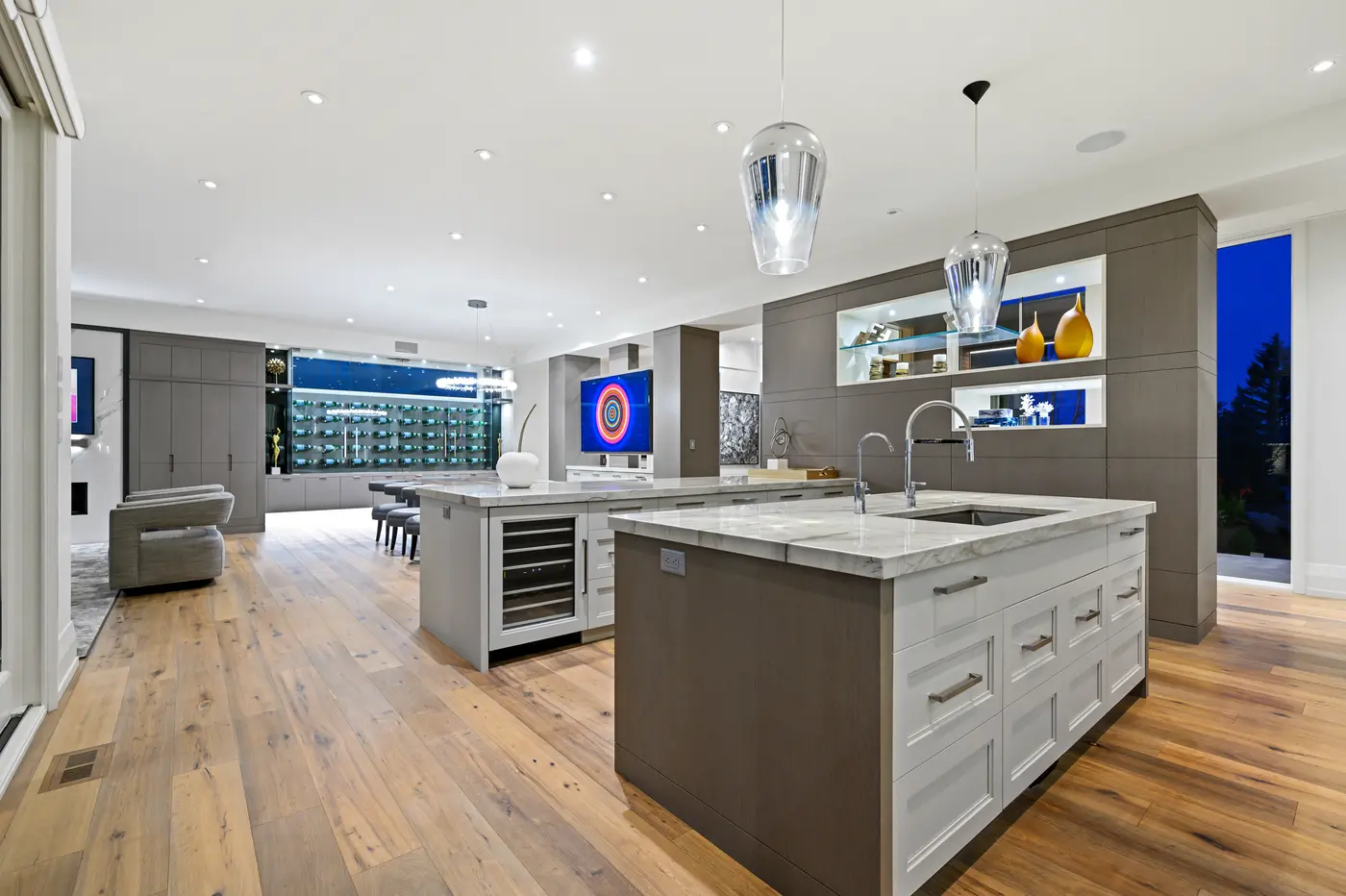 The kitchen's twin islands aligned toward taupe-oak built-ins with lit niches of amber glass, the concentric-ring media art and wine wall carrying colour through the space.