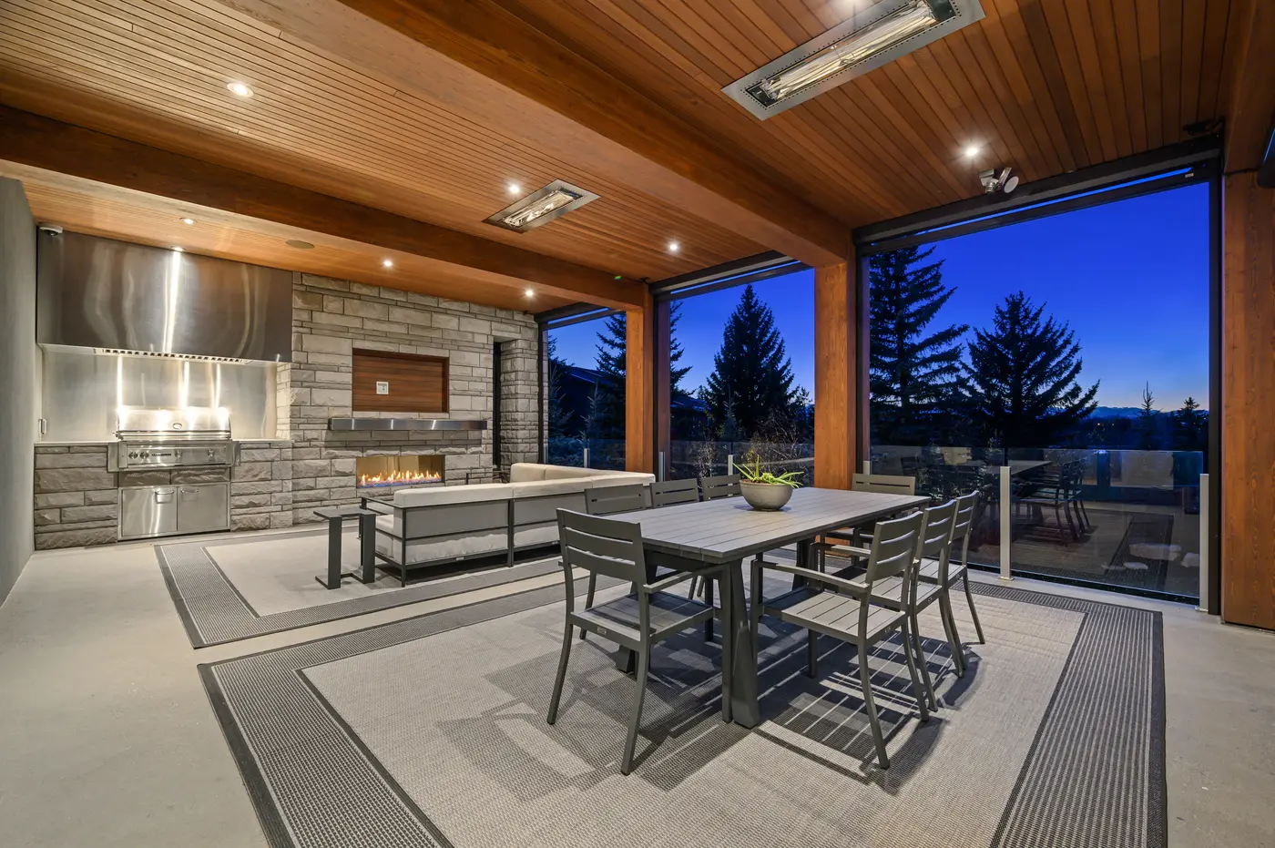 The covered outdoor living room at dusk, infrared heaters set into the cedar ceiling above a built-in grill, stone fireplace wall, and alfresco dining table against glass-railed treetop views.