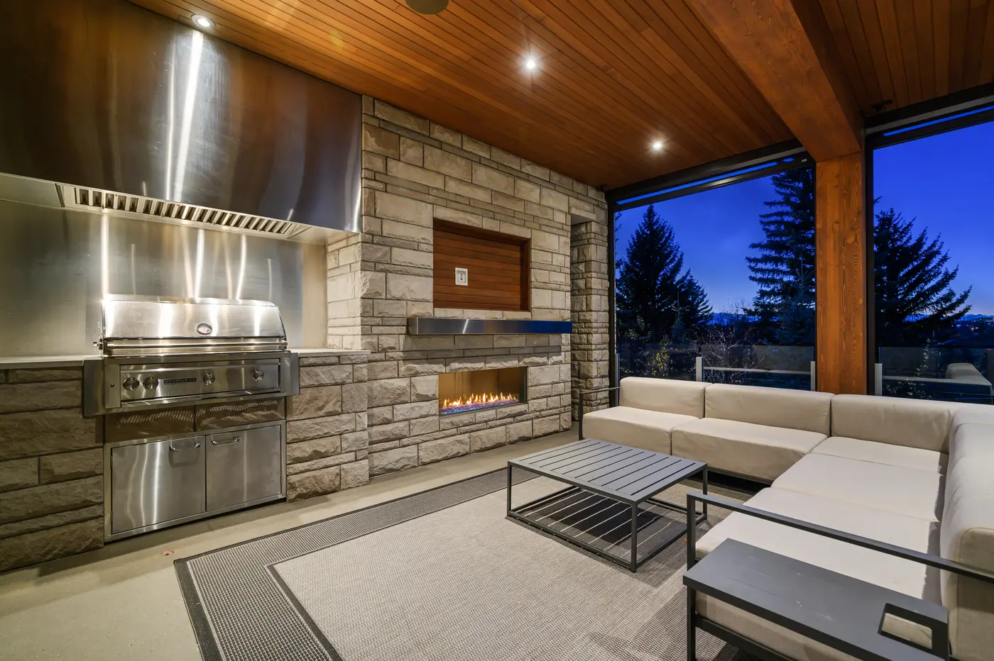 A closer dusk view of the outdoor kitchen's stainless Sedona grill and vent hood beside the limestone fireplace, its linear flame warming a cream sectional under evergreen silhouettes.