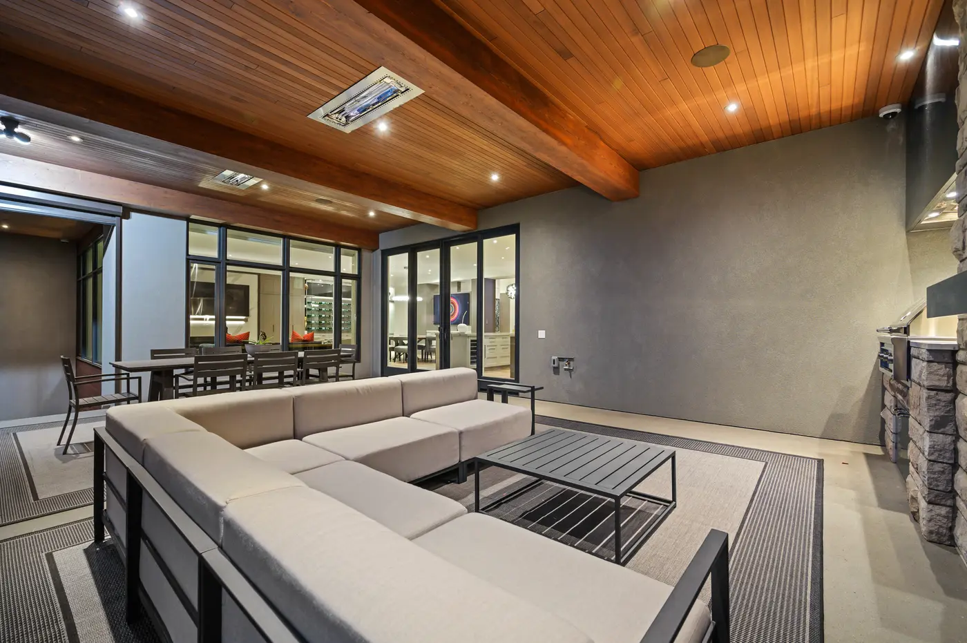 The terrace sectional and slatted coffee table beneath the heated cedar ceiling, glass walls reflecting the lit kitchen and wine display just inside the folding doors.
