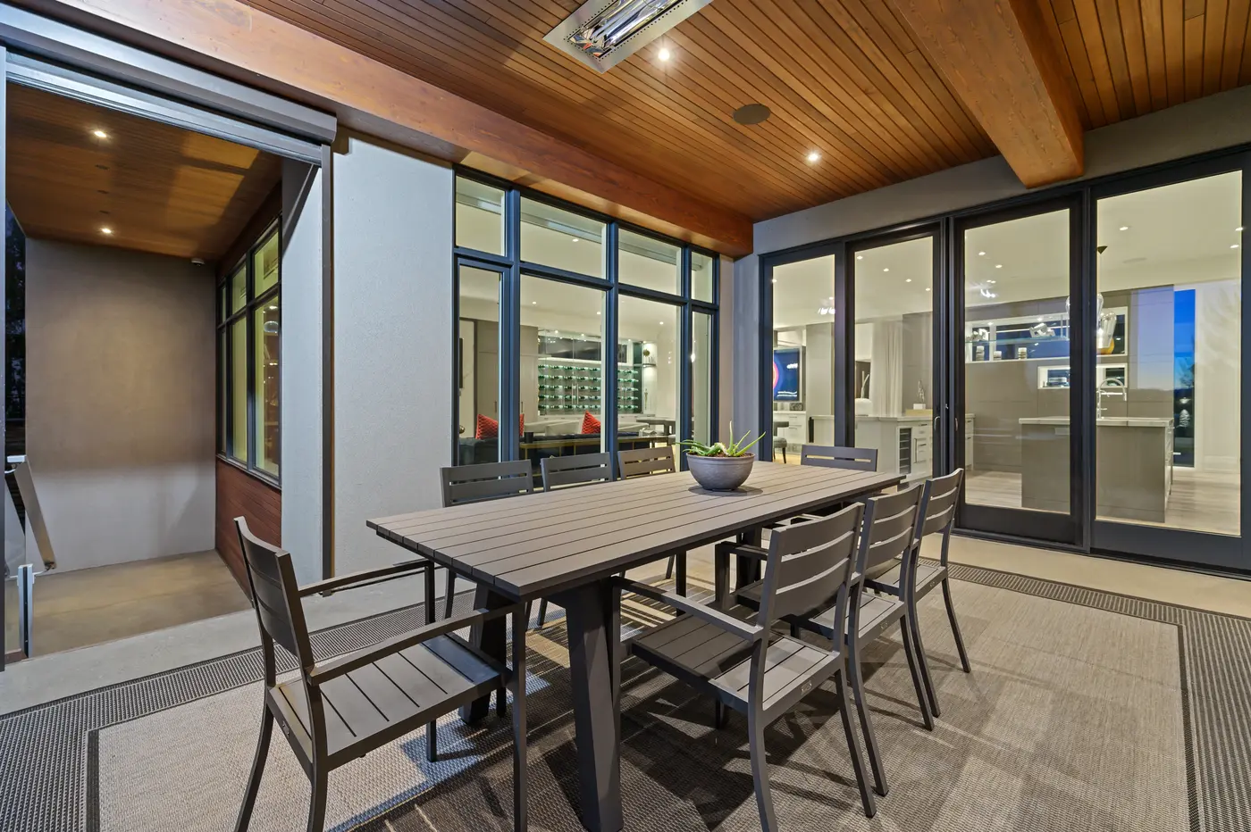 A covered outdoor dining terrace with an eight-seat table beneath a warm cedar-plank ceiling fitted with recessed lighting and infrared heaters, glass doors revealing the illuminated kitchen and wine wall within.