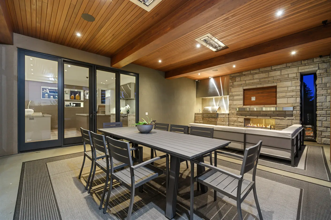The covered outdoor living room at dusk, where a slatted dining table for eight sits before a stone fireplace wall with built-in grill, stainless hood and cushioned lounge seating under a beamed cedar ceiling.