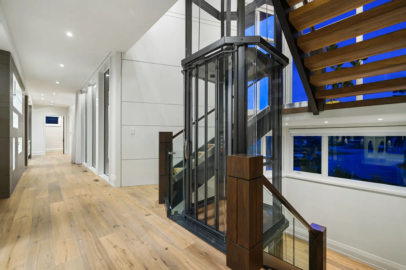 The glass elevator shaft rises beside floating white-oak stair treads and walnut newel posts, framed by twilight-blue windows and a wide-plank oak hallway with gallery millwork.