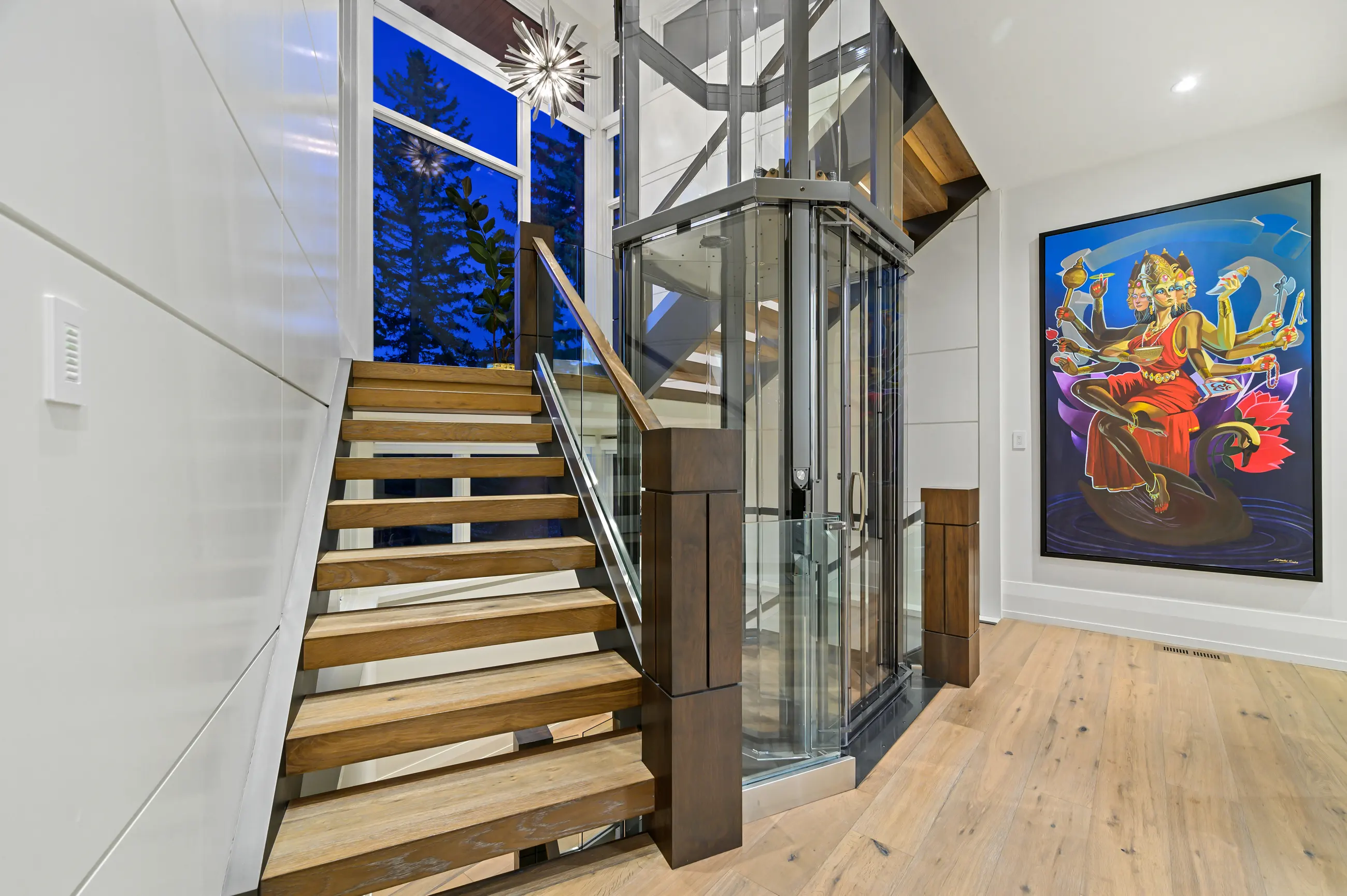 Floating oak stairs with a walnut-capped glass railing climb past the glass elevator cab under a starburst chandelier, with a vivid multi-armed goddess painting anchoring the landing wall.