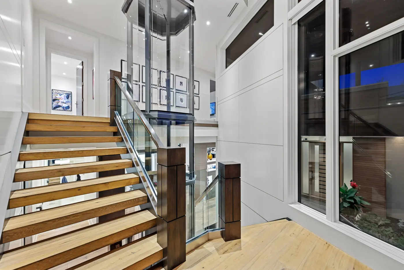 A mid-landing view of the open-riser white-oak staircase wrapping the glass elevator, with a black-framed family photo gallery and twilight glow filtering through the stairwell windows.