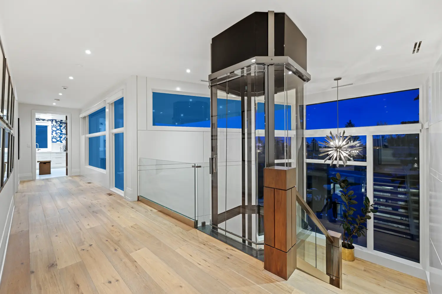 The upper landing at dusk, where the polished glass elevator shaft meets deep-blue clerestory windows, a silver starburst pendant and wide-plank oak floors running toward the laundry room.