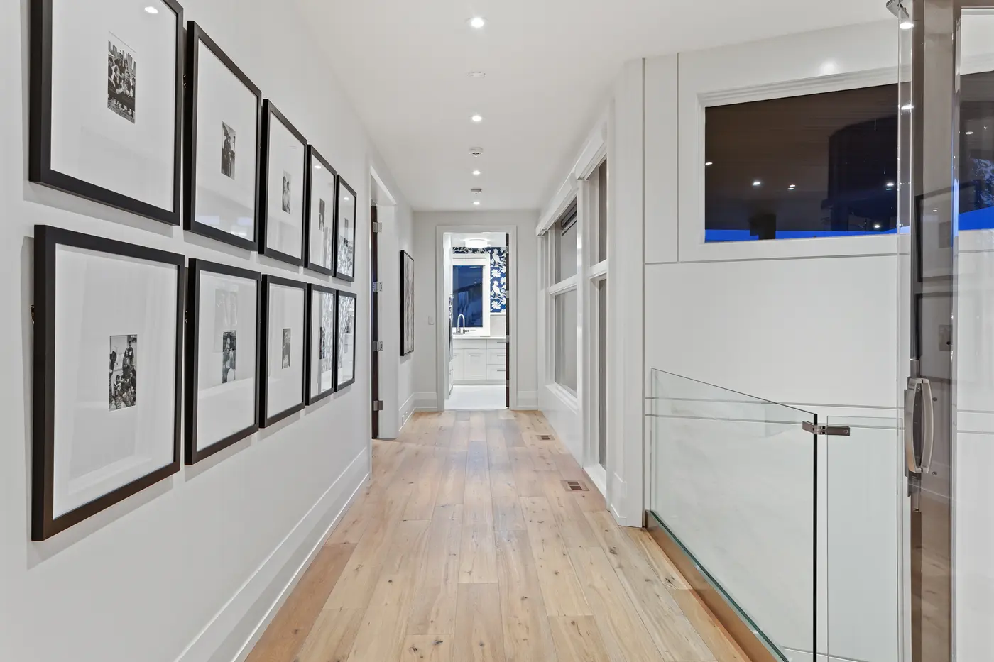 An upper gallery hallway lined with a double row of black-framed family photographs, wide-plank oak flooring and a glass stair rail leading toward a blue floral-papered laundry room.